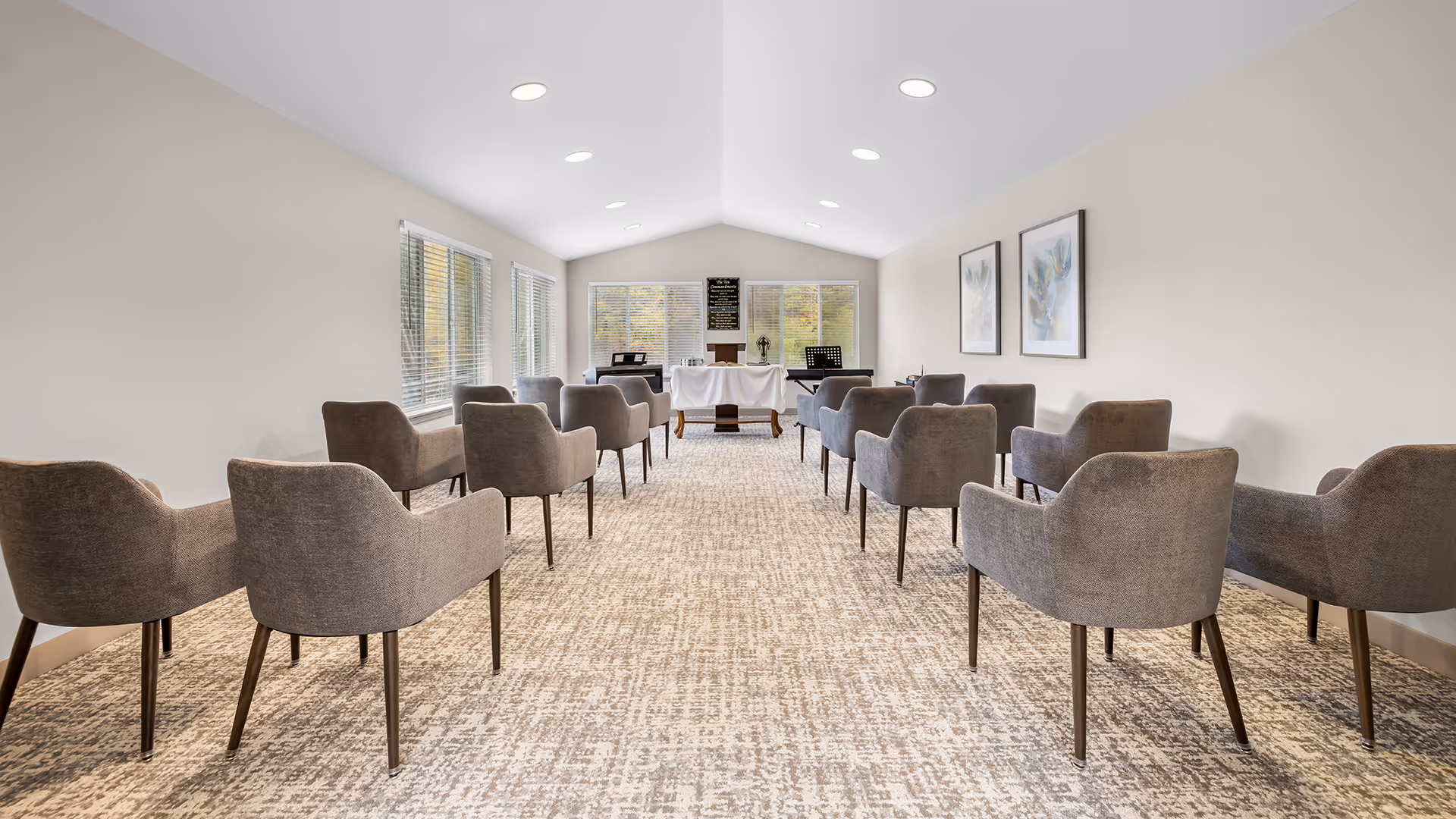 A bright, spacious room with beige walls and carpeted floor, arranged with two rows of gray upholstered chairs facing a small altar table covered with a white cloth. Large windows on the left side let in natural light, and two framed botanical prints hang on the right wall. The room appears to be set up for a small gathering or service.