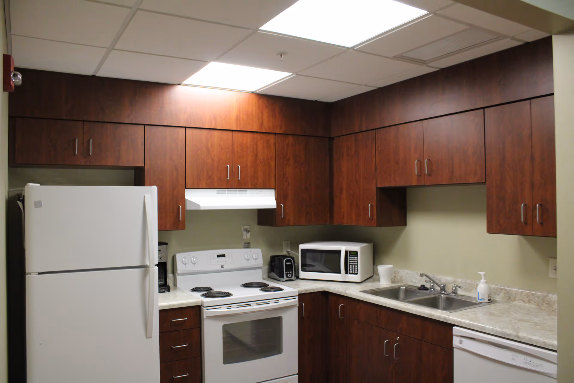A kitchen with wooden cabinets, a white refrigerator, an electric stove with oven, a microwave, a toaster, a double sink, and a dishwasher. The countertop is light-colored, and the ceiling has fluorescent lighting.