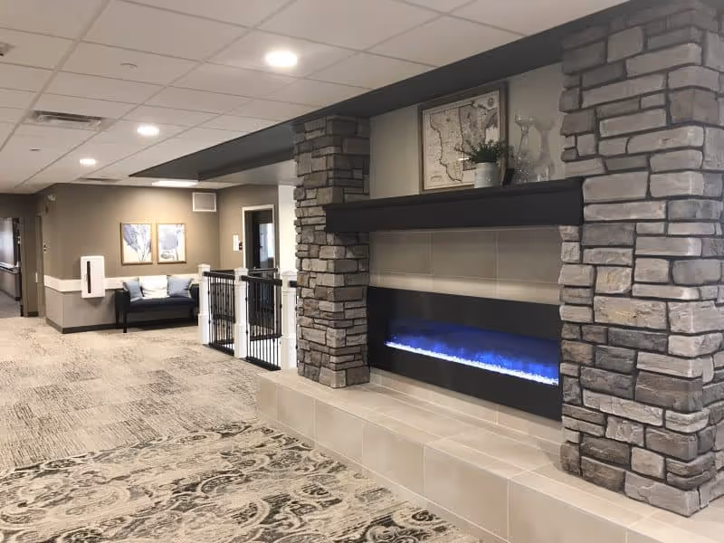 A senior living facility common area with stone pillars, a built-in electric fireplace with blue flames, patterned carpet, and a seating bench in the background.