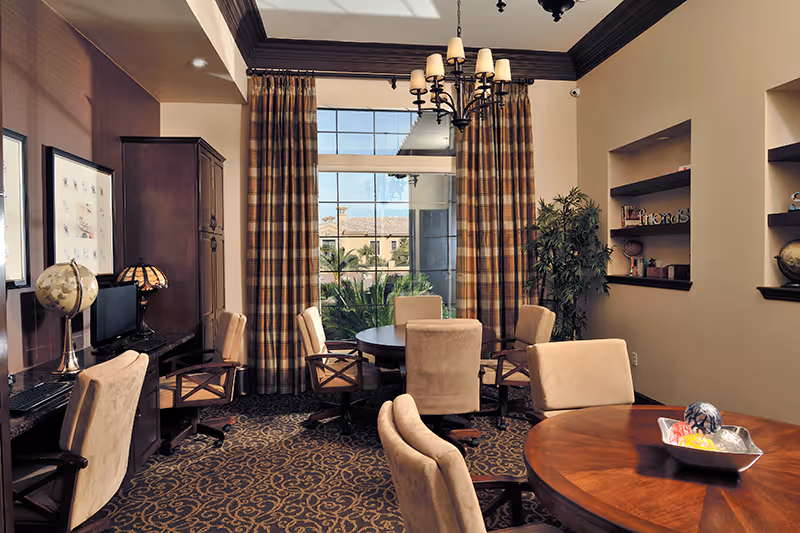 A cozy interior room with round wooden tables surrounded by beige cushioned chairs on wheels. The room features large windows with plaid curtains, a chandelier hanging from the ceiling, a globe on a desk, built-in shelves with decorative items, and a patterned carpet. Outside the window, palm trees and a building are visible.
