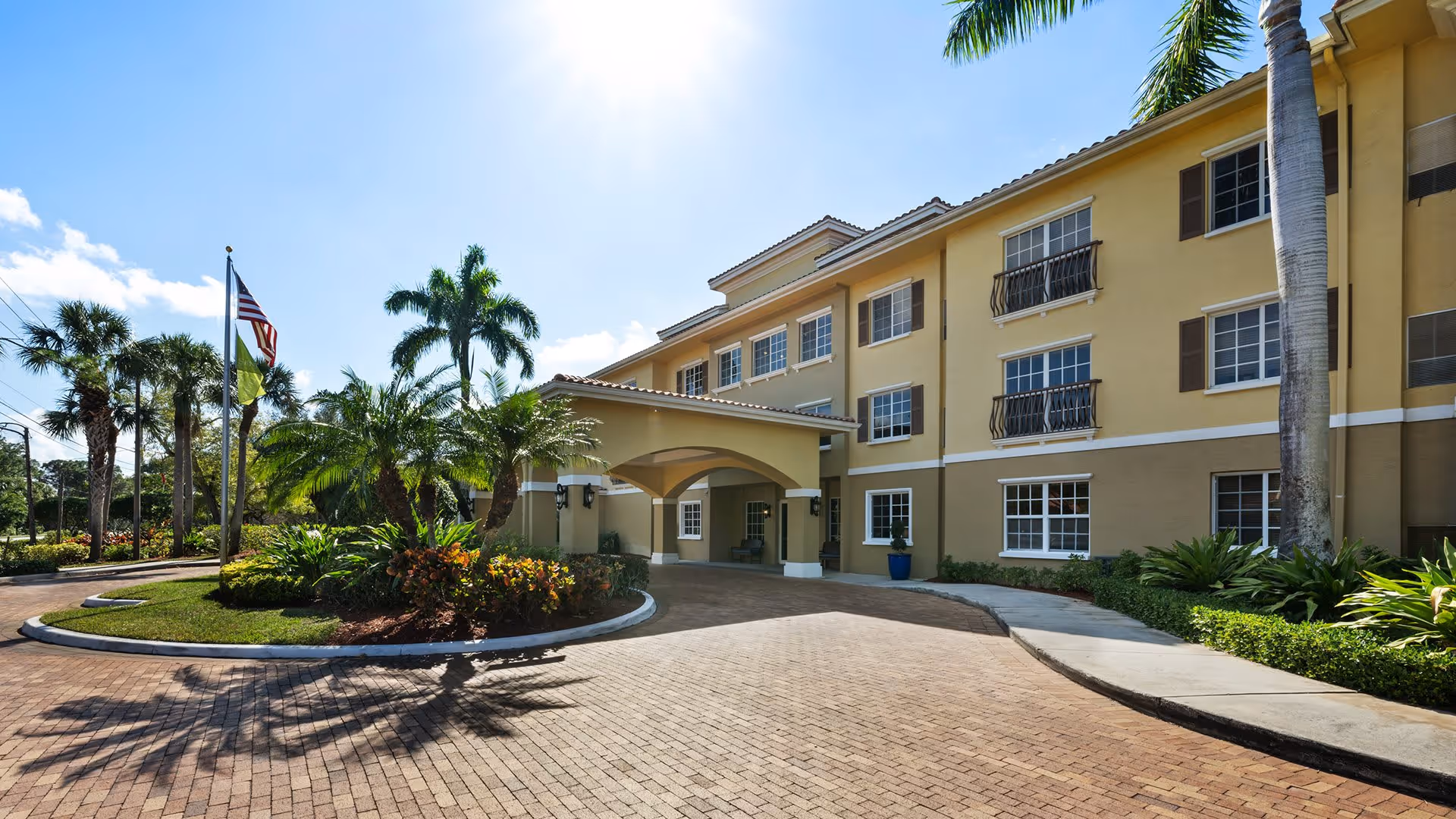 Exterior view of a three-story senior living facility building with a covered entrance driveway, surrounded by palm trees and landscaped greenery under a clear blue sky.
