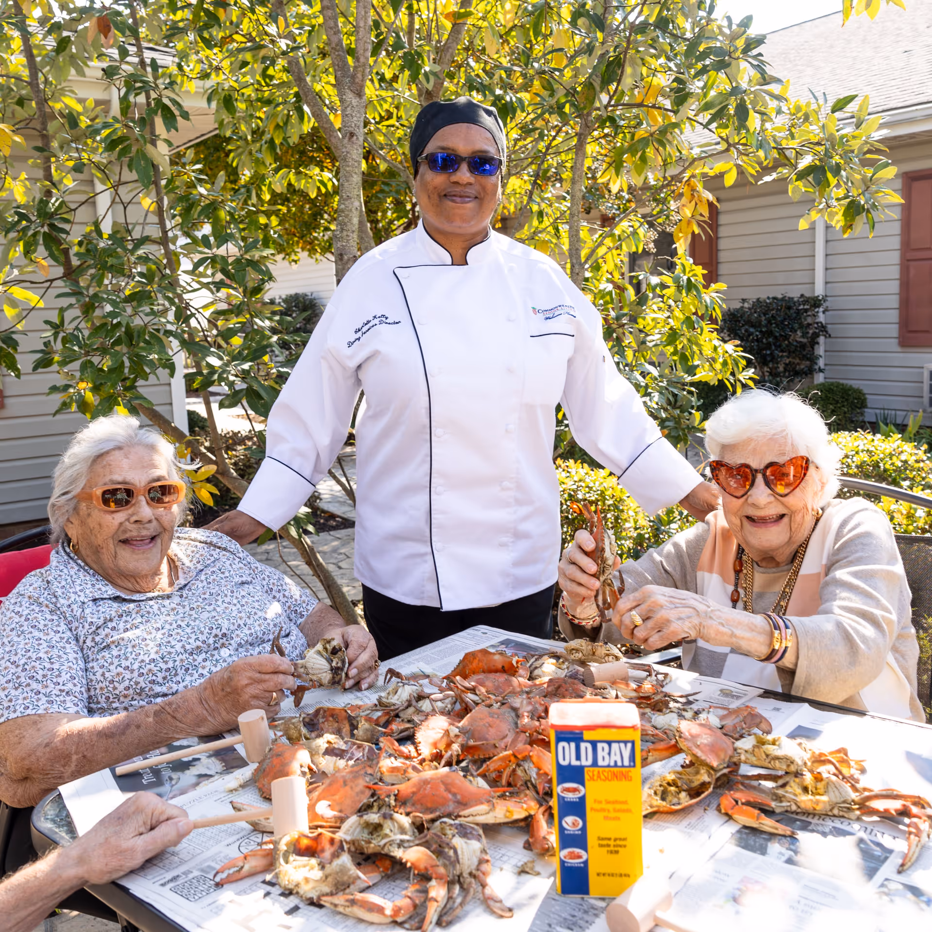 Two elderly women wearing sunglasses sit at a table outdoors enjoying a crab feast with a chef standing behind them. The table is covered with newspapers and a large pile of cooked crabs along with a container of Old Bay seasoning. The setting is a sunny garden area with greenery and a building in the background.