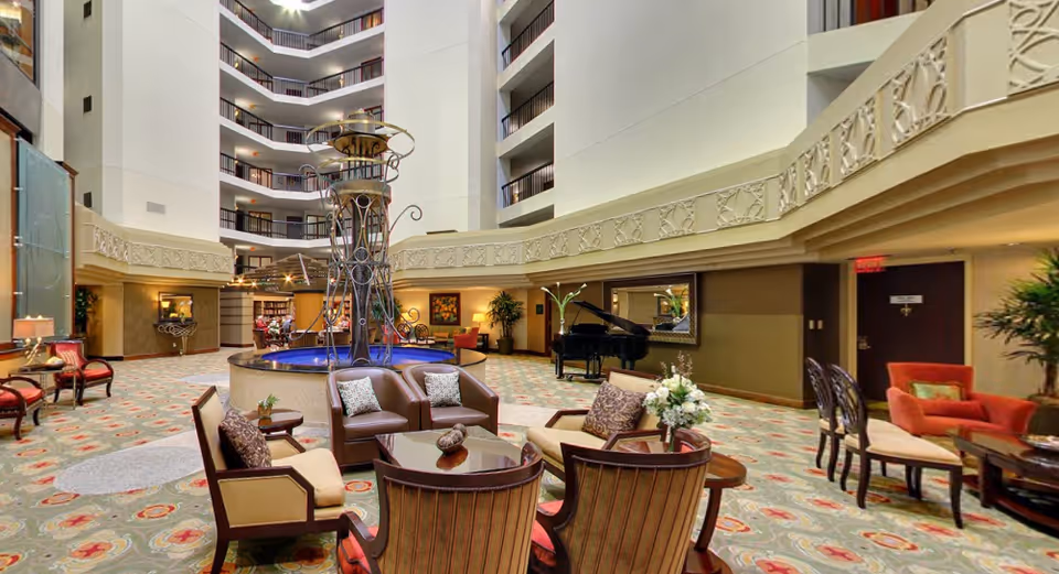 Spacious and elegant interior common area of a senior living facility with a central decorative fountain, comfortable seating arrangements including armchairs and sofas, a grand piano, and multiple floors with balconies overlooking the space.
