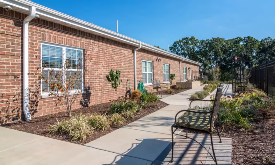 Outdoor walkway alongside a single-story brick building with windows, small landscaped garden beds, and metal benches under a clear blue sky.