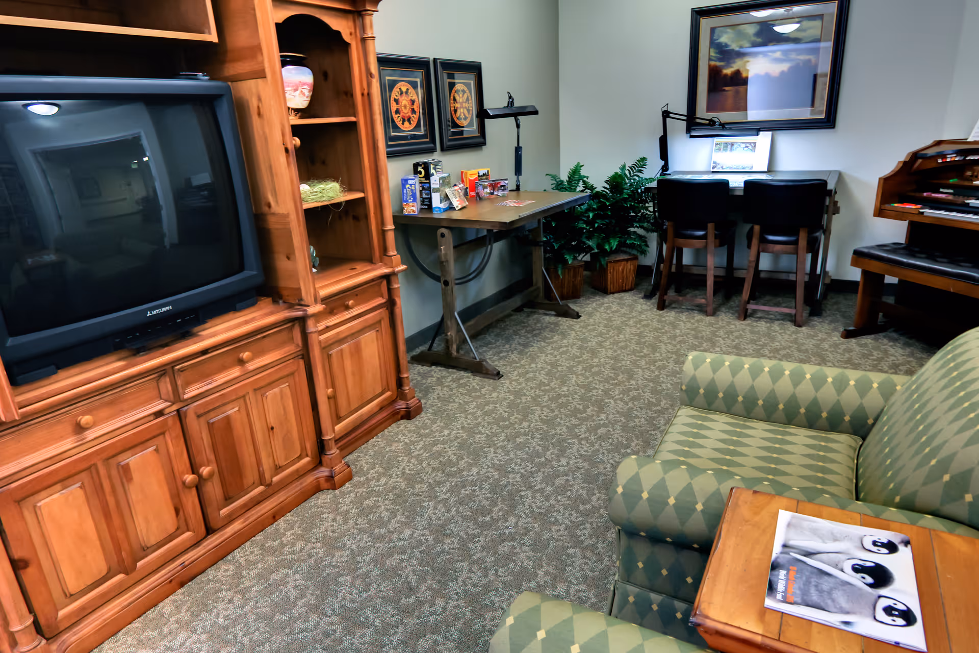 A cozy living room area with a wooden entertainment center holding a large CRT television, a green patterned armchair, a wooden side table with a magazine featuring penguins, a wooden table with various items on it, two chairs at a desk with a lamp, framed artwork on the walls, and a potted plant in the corner.