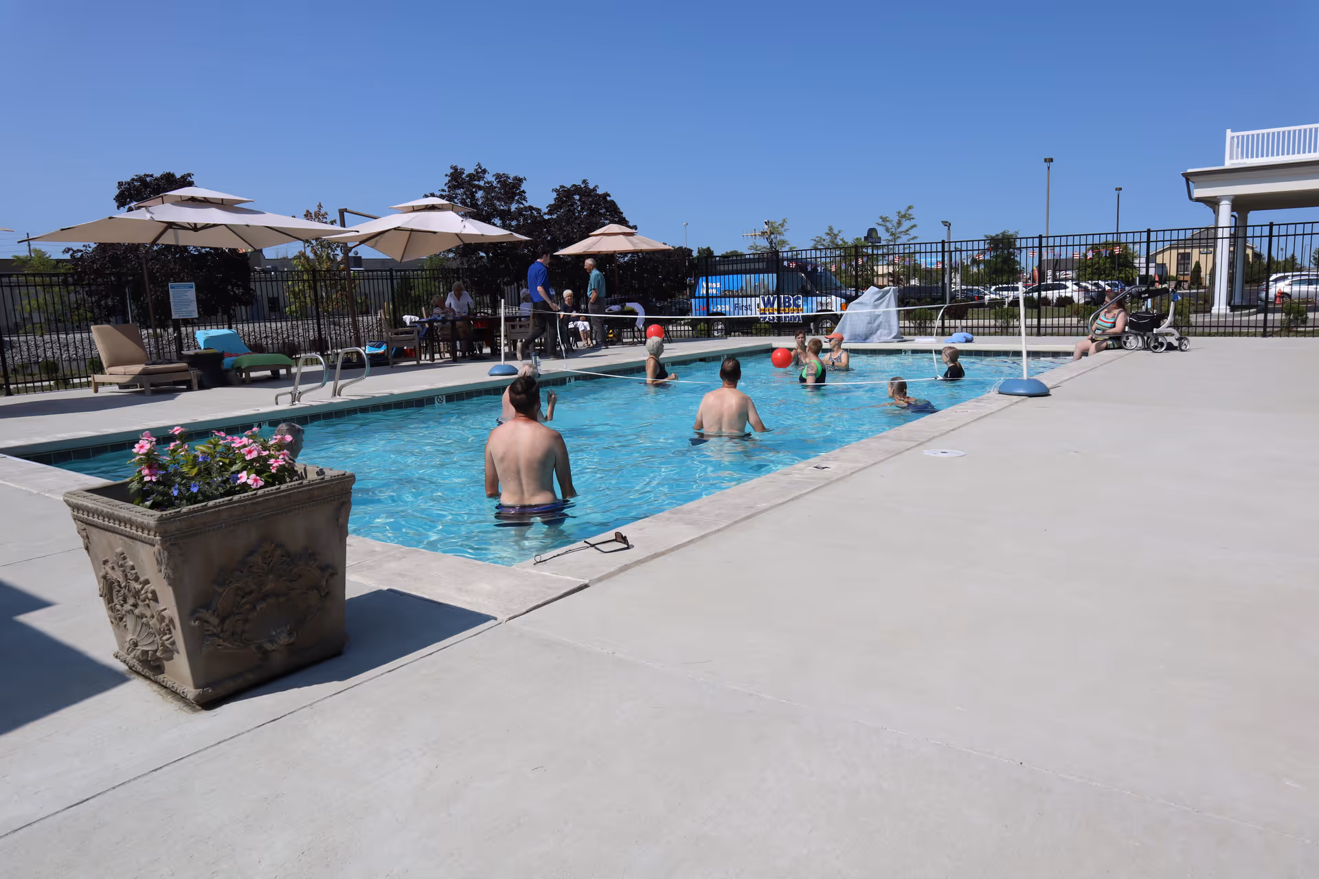 Outdoor swimming pool area with several people in the water playing with a red ball. There are lounge chairs and umbrellas on the pool deck, a large decorative planter with flowers, and a black metal fence surrounding the pool. The sky is clear and blue.