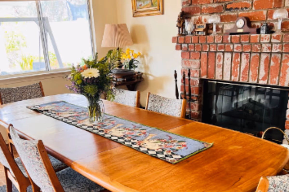 A dining room with a wooden table and six chairs with patterned upholstery. A colorful table runner and a vase with flowers are on the table. There is a brick fireplace with a mantel holding decorative items and a clock. A window with sunlight coming through is on the left side, and a lamp and small table are in the corner.