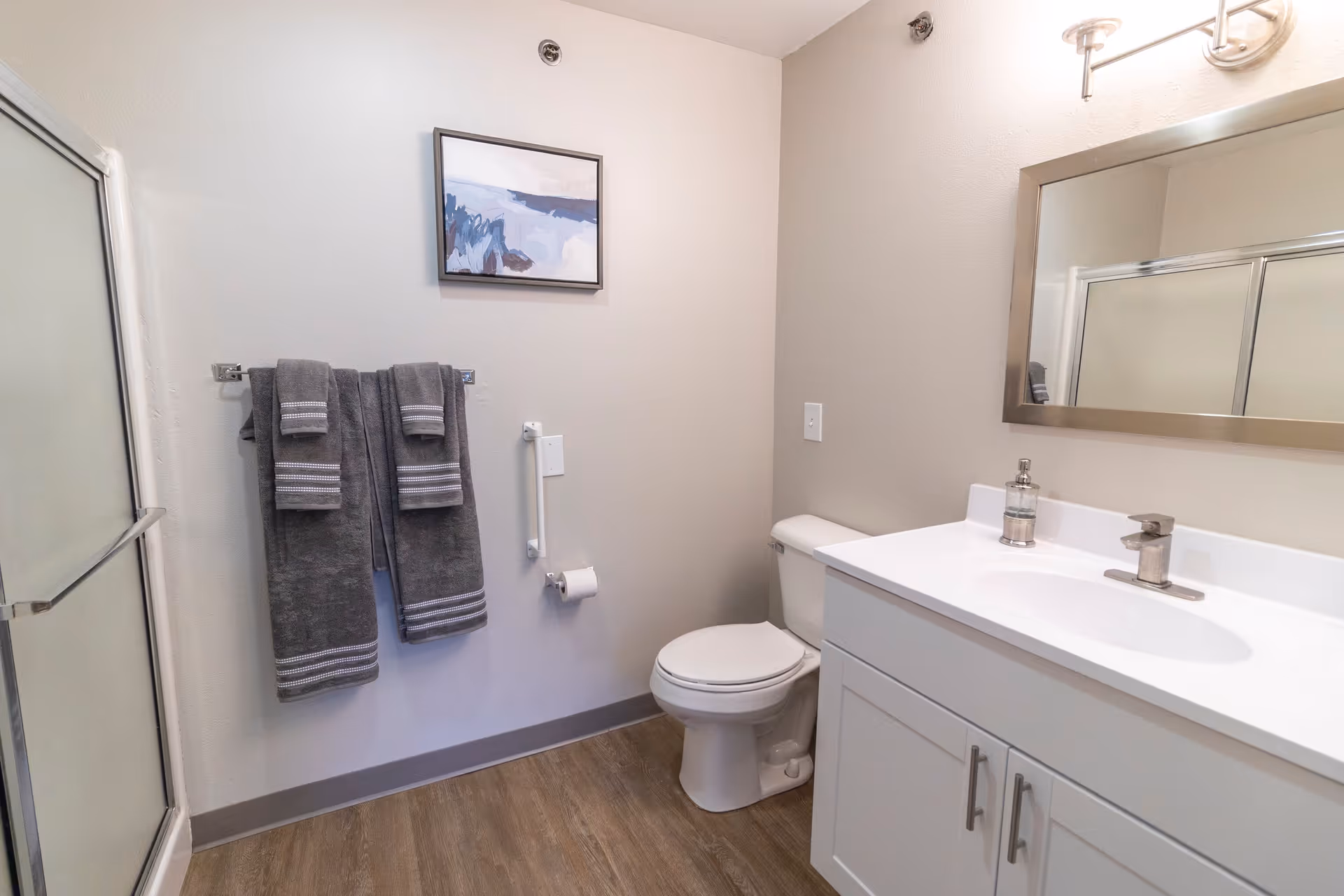 A clean and modern bathroom featuring a white toilet, a white vanity with a sink and faucet, a large rectangular mirror above the sink, a glass-enclosed shower on the left, and three gray towels hanging on a towel rack. There is a small piece of abstract artwork on the wall above the towels, and a toilet paper holder mounted on the wall next to the toilet. The floor has wood-style flooring.