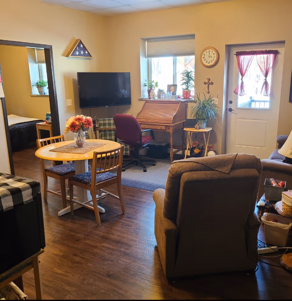 Interior living area with a recliner, small dining table, wall-mounted TV, desk, and a door with a window.