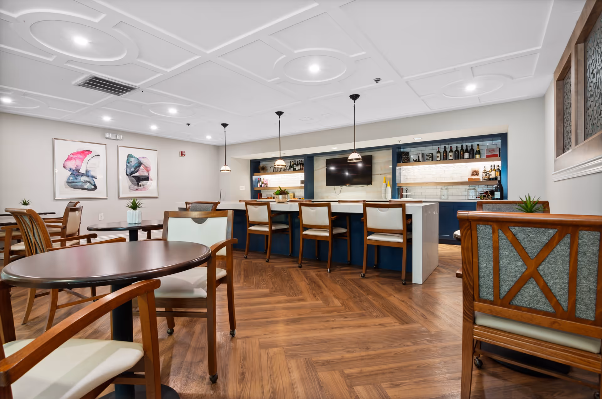 A modern common area with wooden flooring and multiple round tables with chairs. The back wall features a bar counter with four chairs, pendant lights hanging above, and shelves stocked with bottles and decorative items. Two abstract paintings hang on the left wall, and small potted plants are placed on some tables.