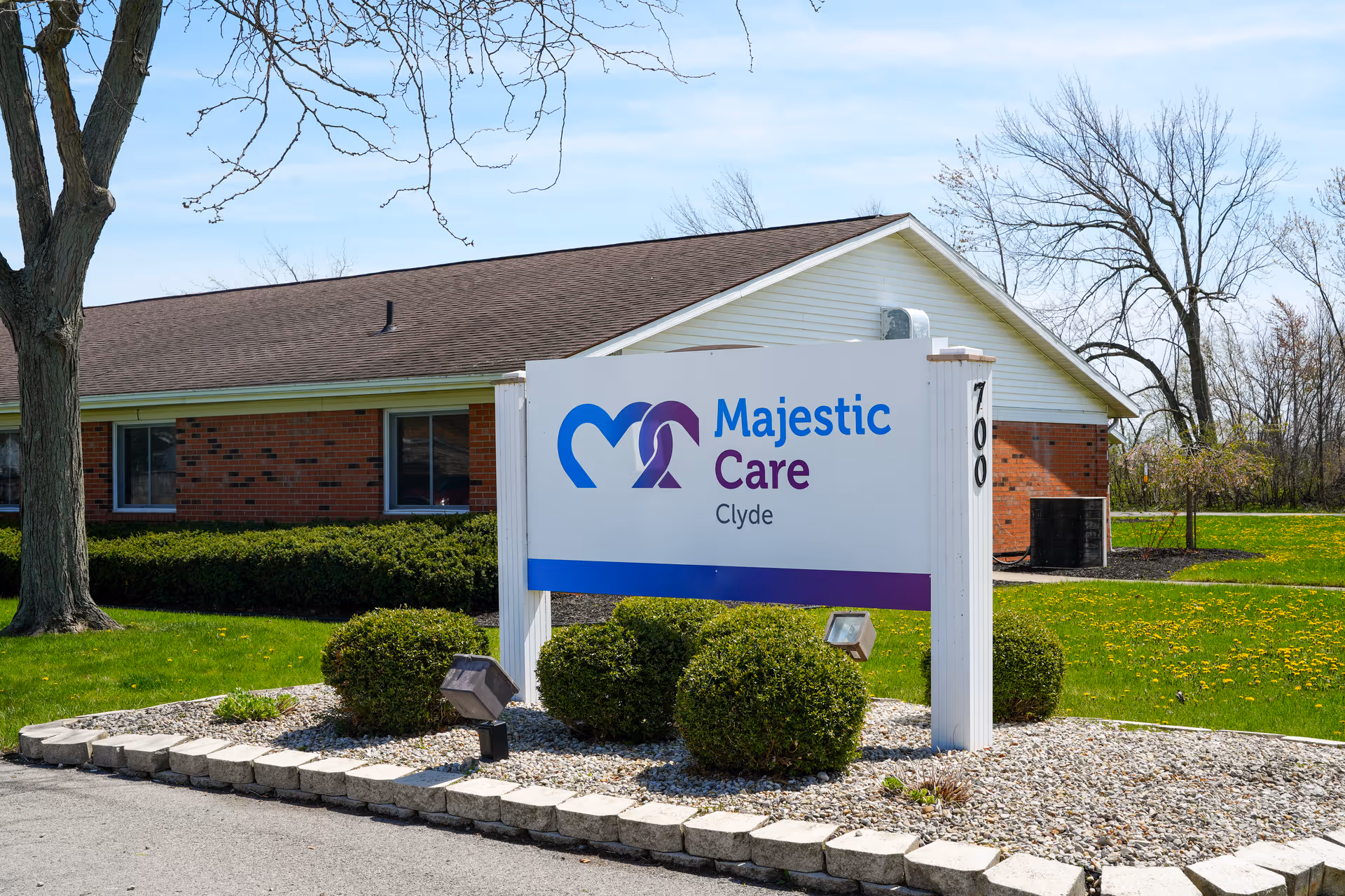 Outdoor view of the Majestic Care of Clyde facility sign with a brick building and green lawn in the background under a blue sky.