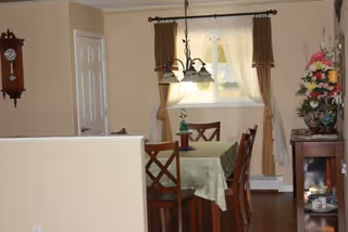 A dining area with a wooden table covered with a green tablecloth and four wooden chairs. A chandelier with five lights hangs above the table. There is a window with sheer white curtains and brown drapes behind the table. To the right, there is a wooden cabinet with a floral arrangement on top and some decorative items inside. A wall clock is mounted on the left wall near a closed white door.