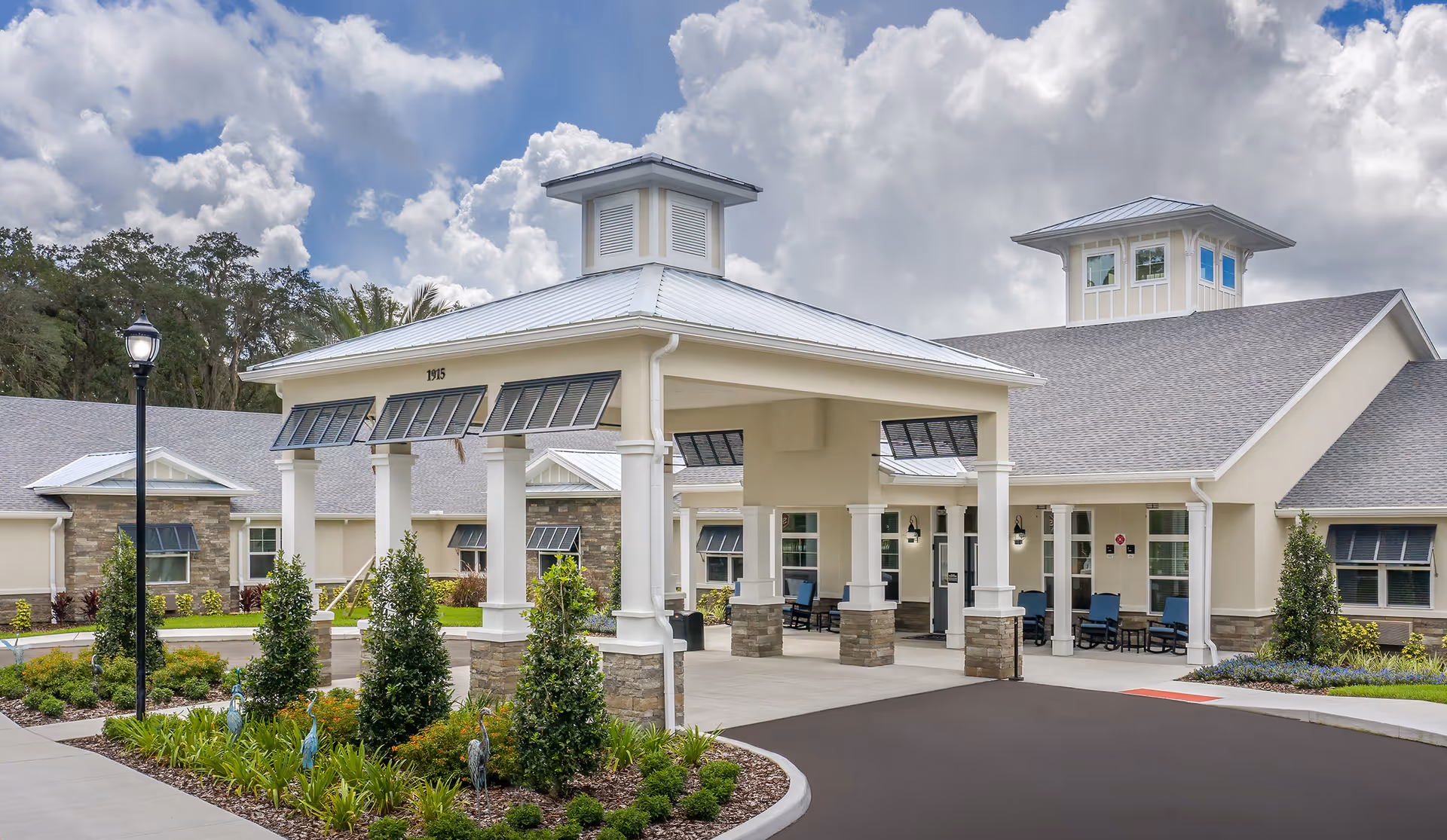 Exterior front entrance of a senior living community with a covered porte-cochere, landscaped gardens, and outdoor seating.