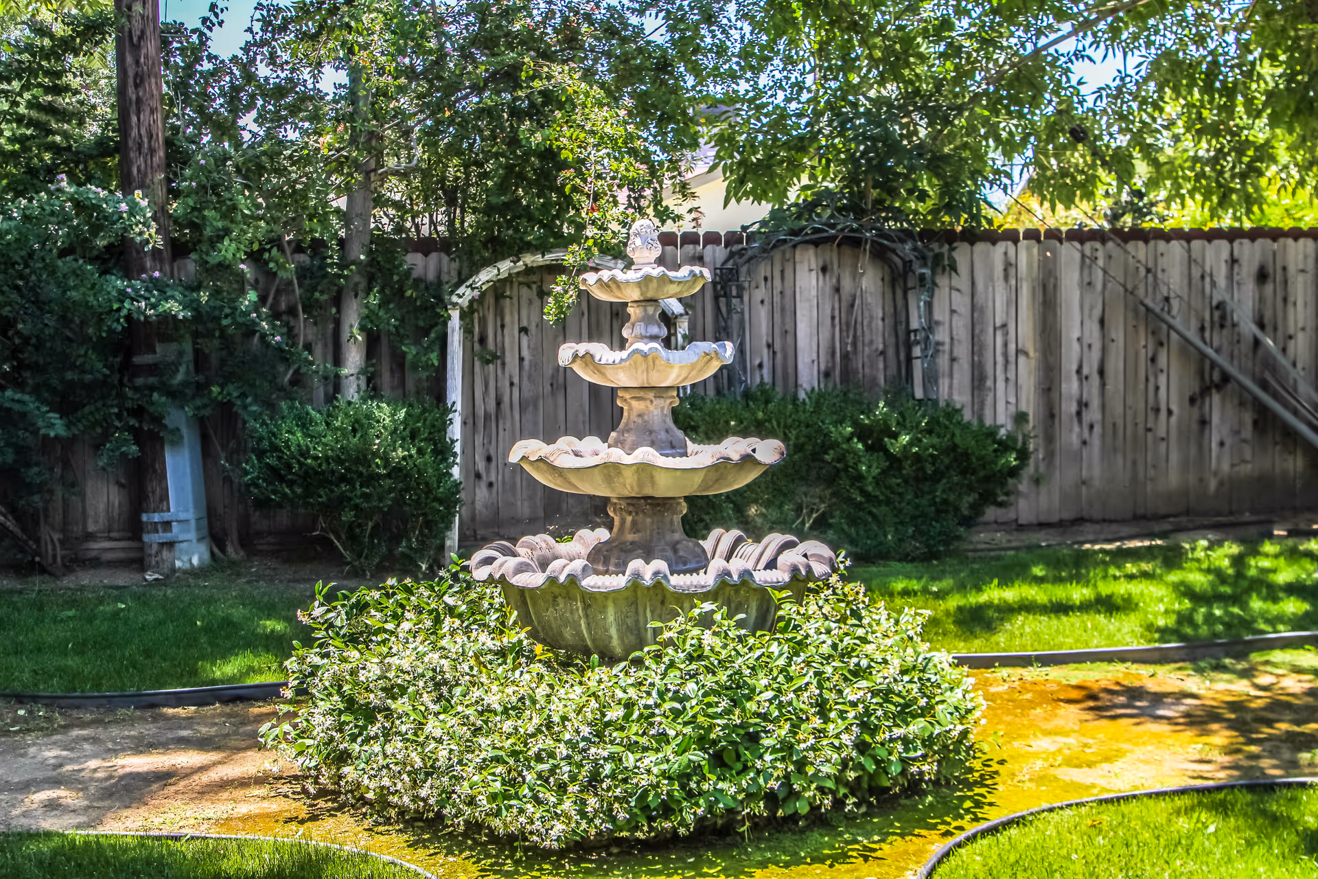 A multi-tiered stone fountain surrounded by green bushes and grass in a garden area with a wooden fence and trees in the background.