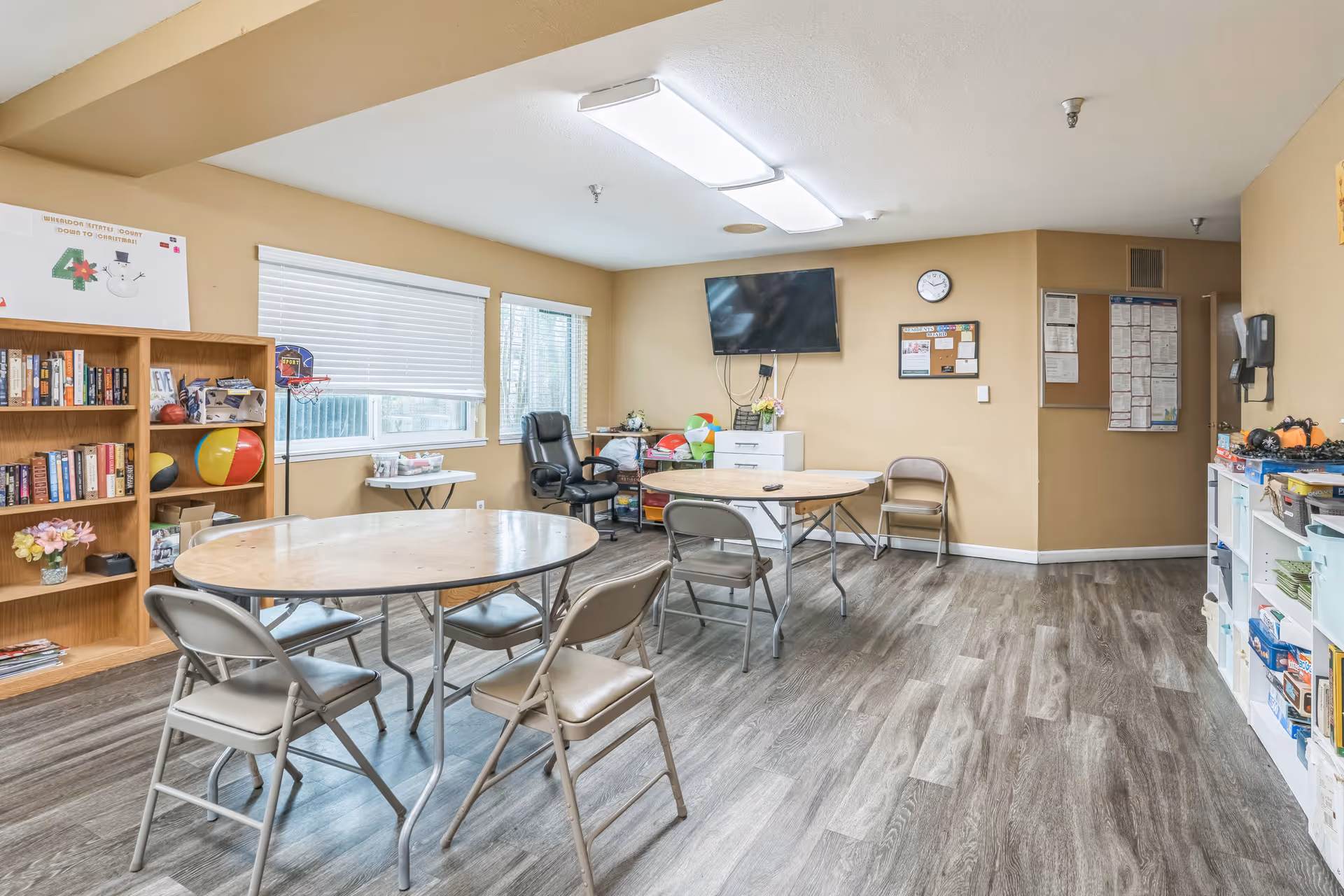 A bright activity room with two round tables surrounded by folding chairs, a bookshelf filled with books and games, a TV mounted on the wall, and windows with blinds letting in natural light. The room has wood-look flooring and beige walls with various bulletin boards and decorations.