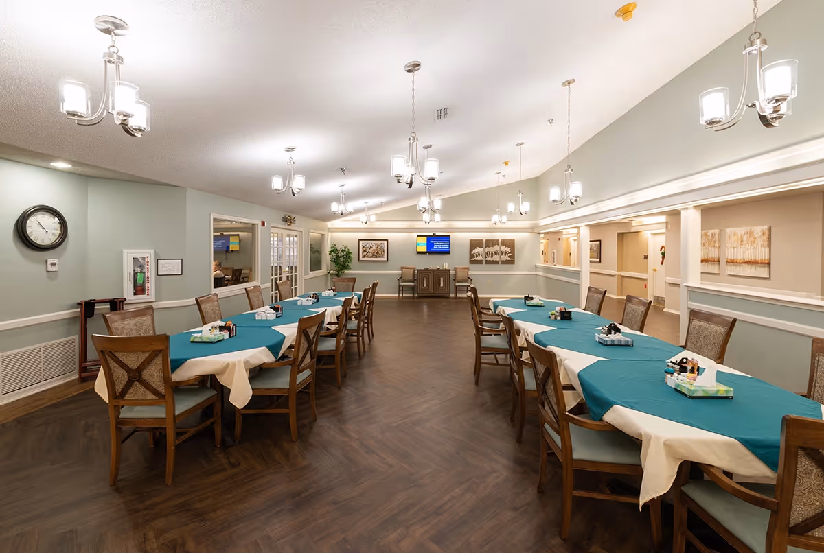 A spacious dining room with two long tables covered with teal and white tablecloths, surrounded by wooden chairs with cushioned seats. The room has multiple modern chandeliers hanging from the ceiling, light green walls, and dark wood flooring. There is a clock on the left wall, a fire extinguisher cabinet, framed artwork, and a flat screen TV mounted on the far wall.