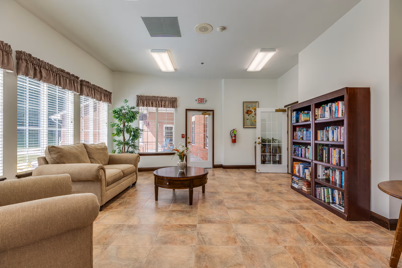 A bright and spacious common area with large windows covered by brown valances, a beige sofa and armchair, a round wooden coffee table with a vase of flowers, a tall green plant, and a wooden bookshelf filled with books. There is a door with a glass panel and an exit sign above it, a fire extinguisher mounted on the wall, and a framed picture hanging next to the door.