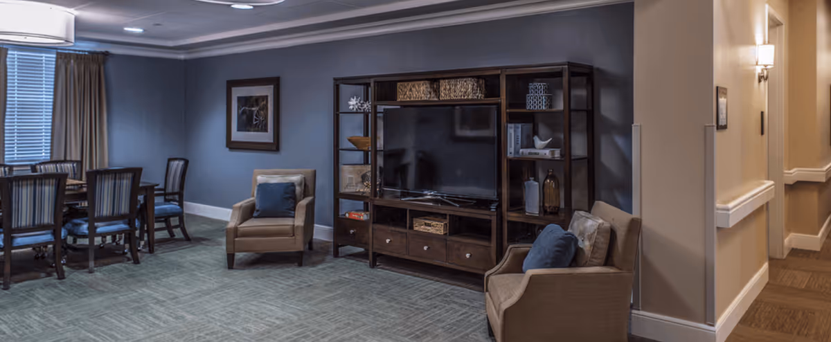 A cozy common area featuring a large flat-screen TV on a wooden entertainment center with shelves holding decorative items. Two beige armchairs with blue cushions are positioned near the TV. To the left, there is a dining table with six chairs, and a window with closed blinds and beige curtains. The walls are painted gray and beige, with a framed picture hanging on the wall. The floor is carpeted, and there is a hallway visible on the right side of the image.