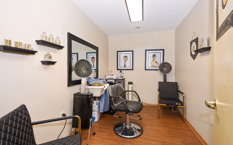 Small salon room with a styling chair, hooded hair dryer, mirror, shelving, and framed portraits on the wall.
