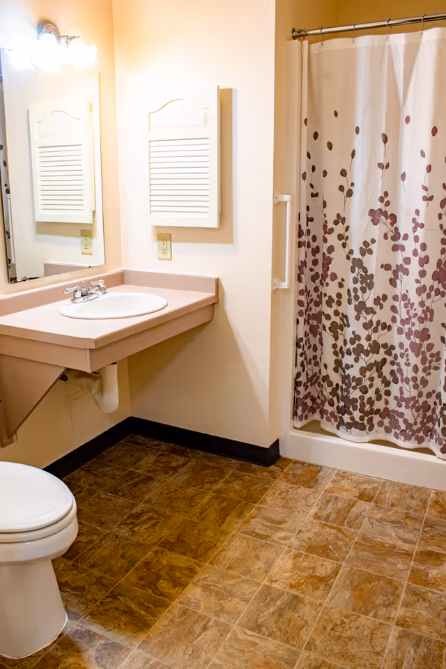 A clean bathroom with a white toilet, a beige countertop with a built-in sink, a large mirror above the sink, a wall-mounted cabinet with louvered doors, and a shower with a white curtain featuring a pattern of brown and purple leaves. The floor has brown tile, and the walls are painted light beige.