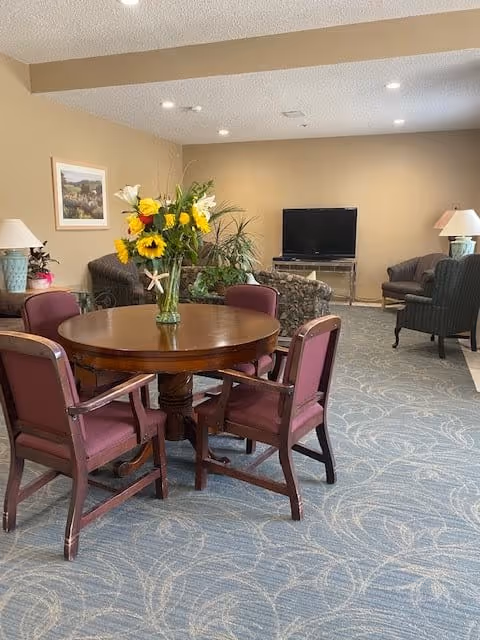 A cozy living room area with a round wooden table surrounded by four maroon cushioned chairs. A vase with a colorful bouquet of flowers, including sunflowers and lilies, is placed on the table. In the background, there are several upholstered armchairs and a flat-screen TV on a stand against a beige wall. The room has a patterned blue carpet and soft lighting from ceiling fixtures and table lamps.