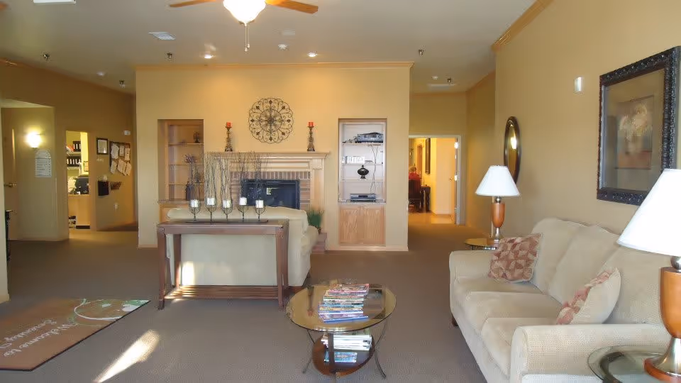 A cozy living room in an assisted living facility with beige walls and carpet. The room features a beige sofa with patterned cushions, a glass coffee table with magazines, a wooden table behind the sofa with decorative candle holders, a fireplace with built-in shelves on either side, and a ceiling fan with a light. There are lamps on side tables and framed artwork on the walls. A hallway leads to other rooms in the background.