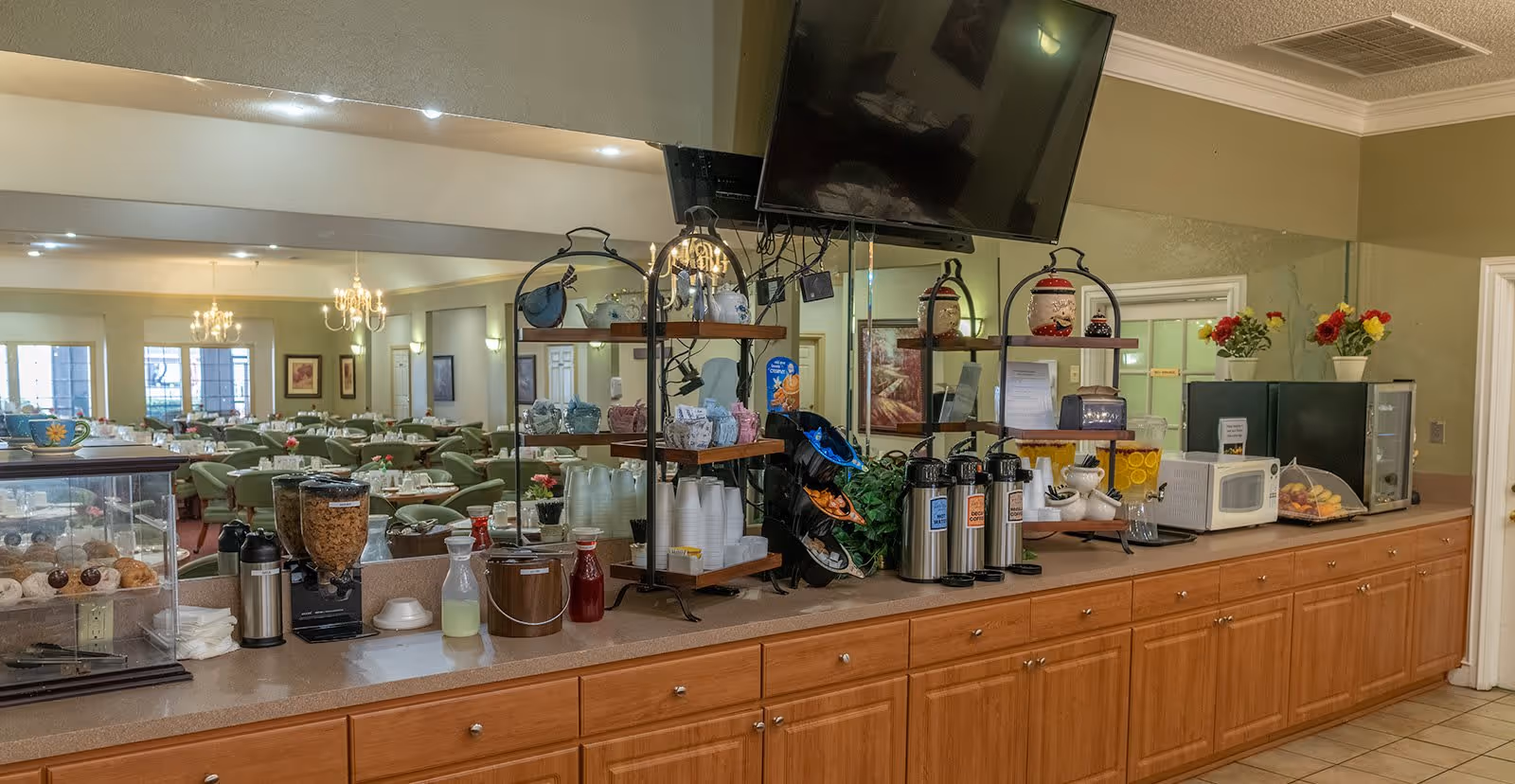 A long countertop in a dining area with various breakfast items including cereal dispensers, coffee pots, cups, a microwave, and a fruit tray. Behind the counter is a large mirror reflecting a dining room with multiple tables and green chairs, each table set with plates and flowers. A flat-screen TV is mounted above the counter.