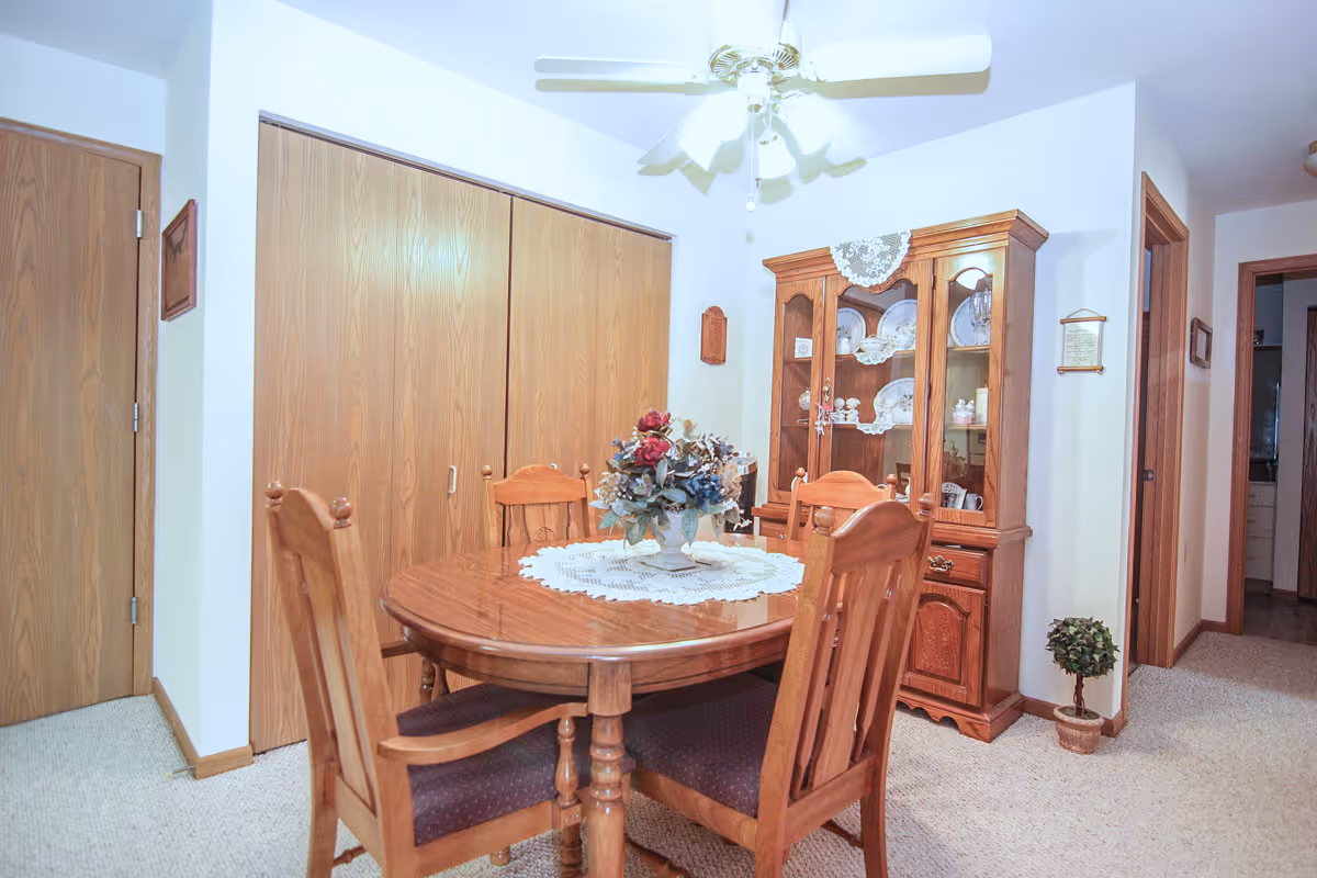 Carpeted dining area with a round wooden table and chairs, a china cabinet, closet doors, and a ceiling fan.