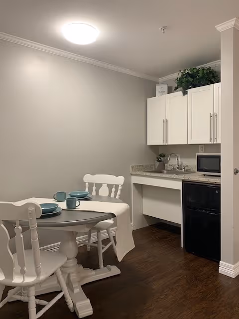 Small dining area with a round white table set with blue plates and mugs, accompanied by two white wooden chairs. Adjacent to the dining area is a compact kitchenette with white cabinets, a granite countertop, a sink, a microwave, and a small black refrigerator. The floor is dark wood, and the walls are light gray.