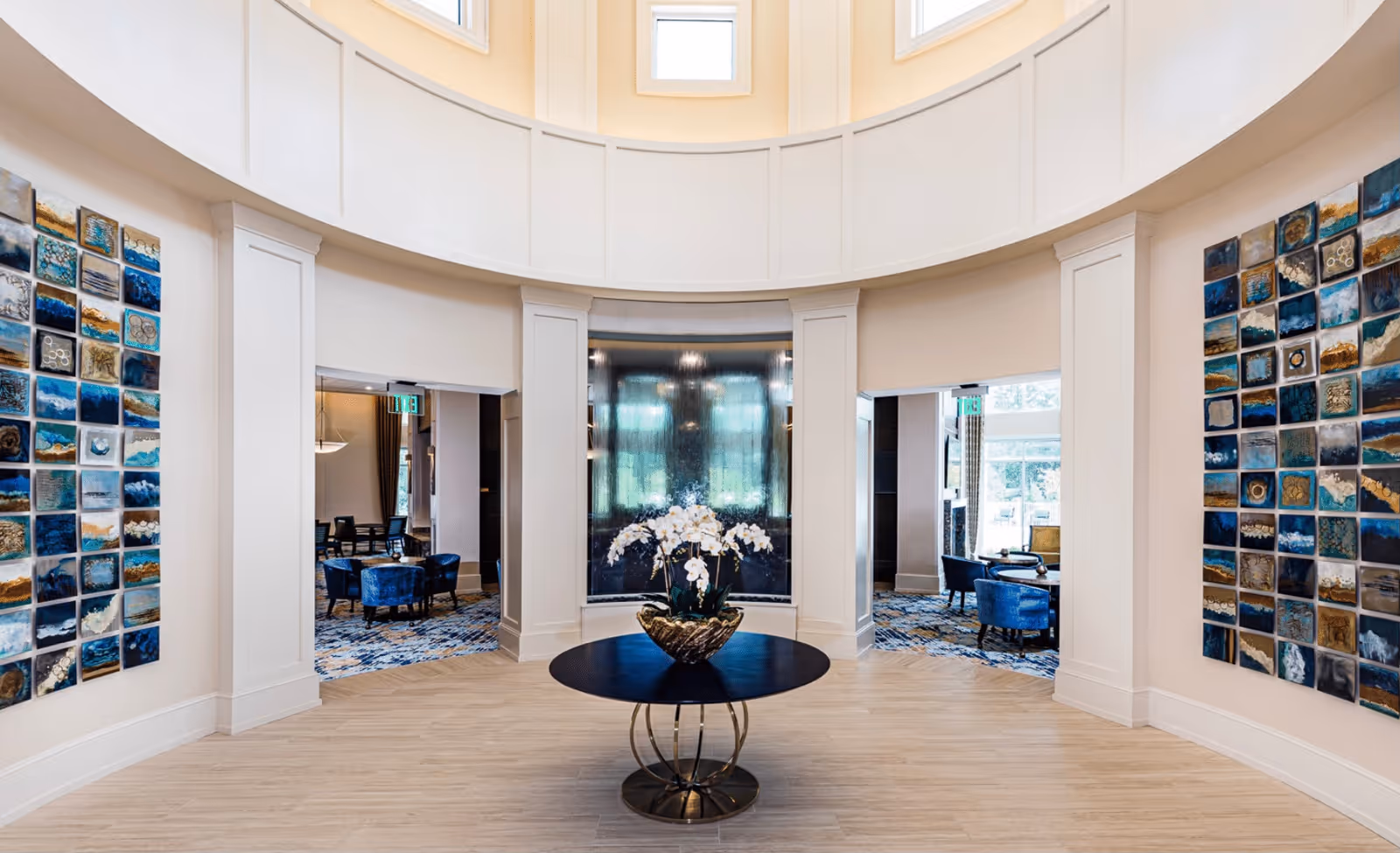 A bright, spacious circular interior area with a round black table in the center holding a decorative vase with white flowers. The walls are adorned with colorful square art pieces on both sides. Two open doorways lead to rooms furnished with blue chairs and tables, with large windows allowing natural light to fill the space.