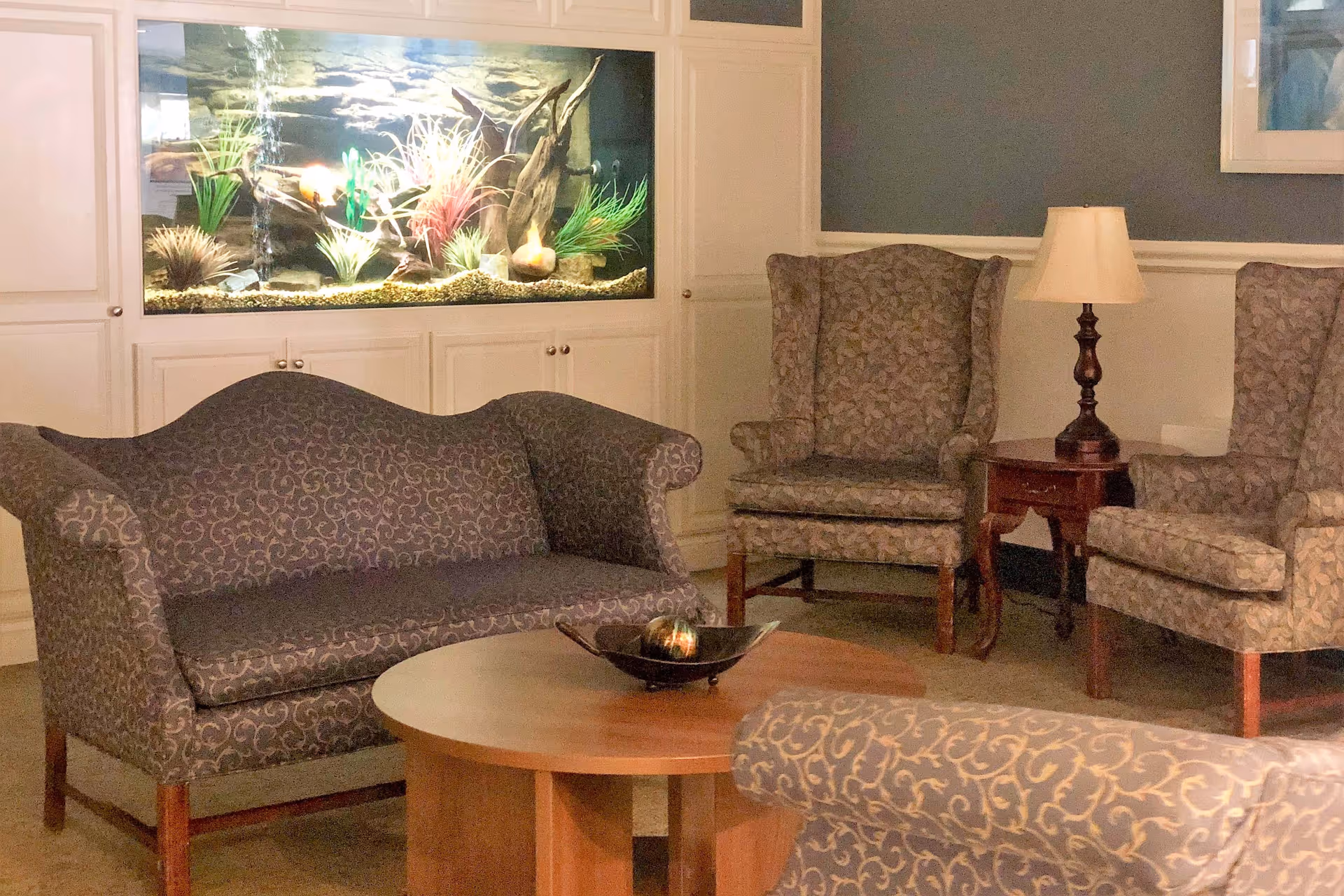 A cozy sitting area with patterned sofa and armchairs around a wooden coffee table and a built-in aquarium in the wall.