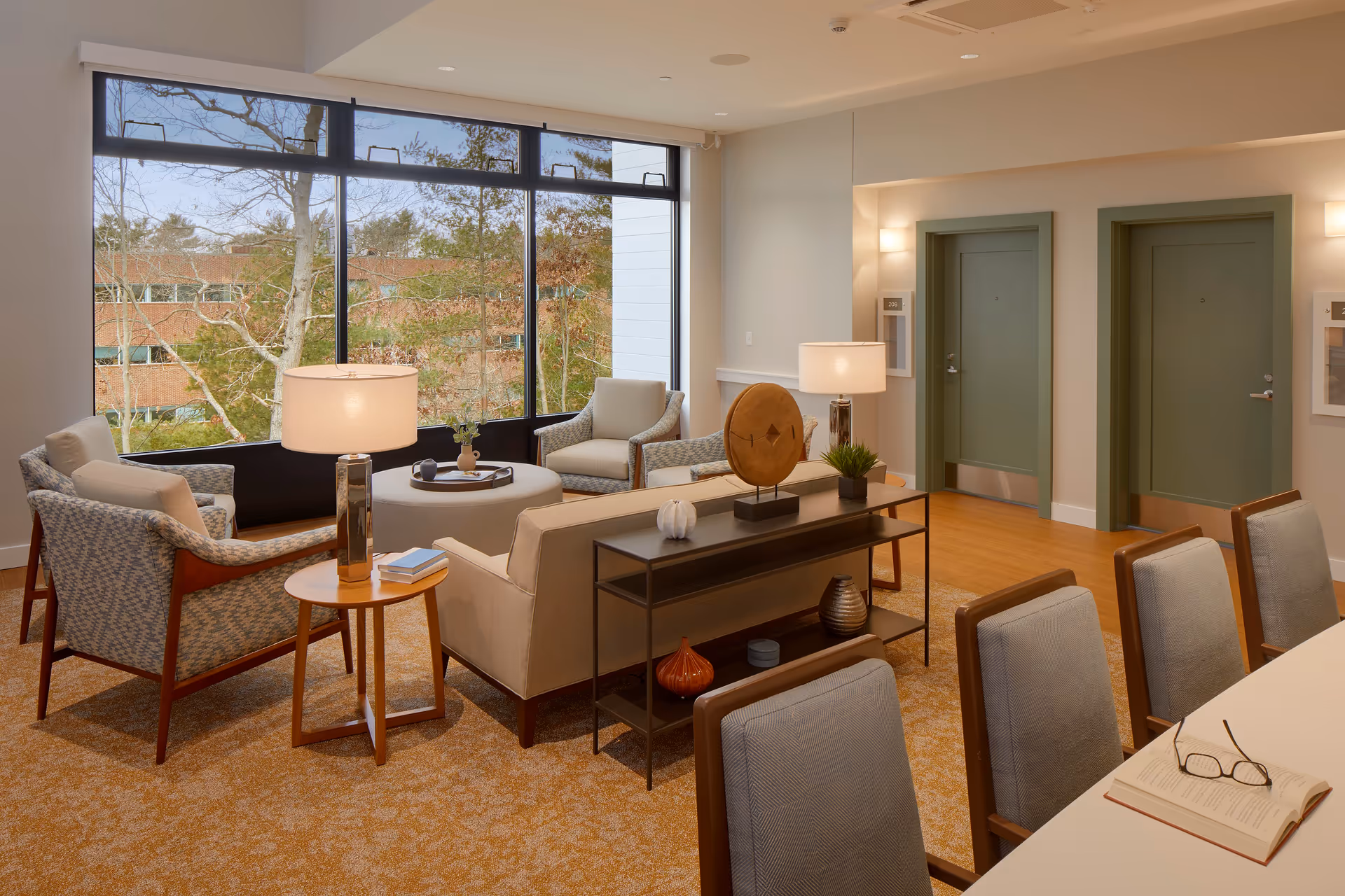 Well-lit communal lounge with sofas, armchairs, side tables, lamps, and a large window overlooking trees.