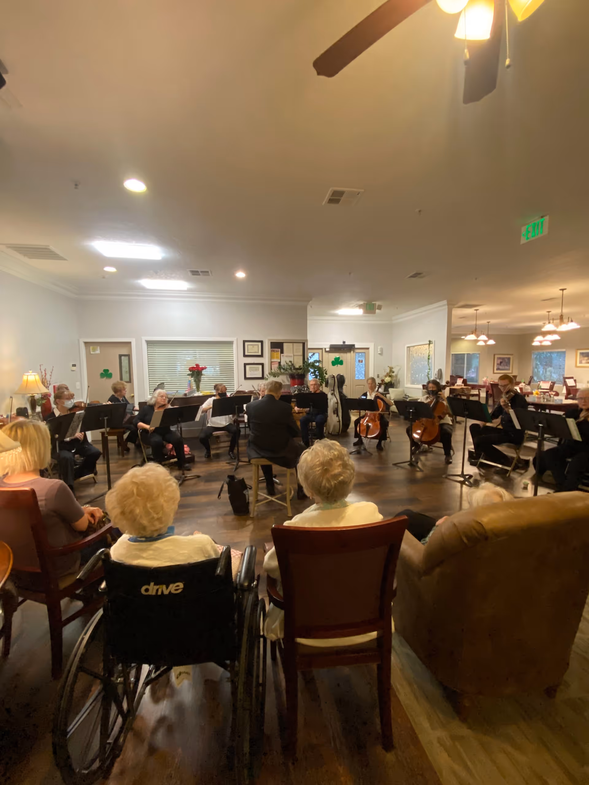 A group of elderly people seated in chairs and a wheelchair watching a small orchestra performing in a well-lit common room with wooden floors and ceiling fans. The musicians are playing string instruments and are seated in a semicircle facing the audience.