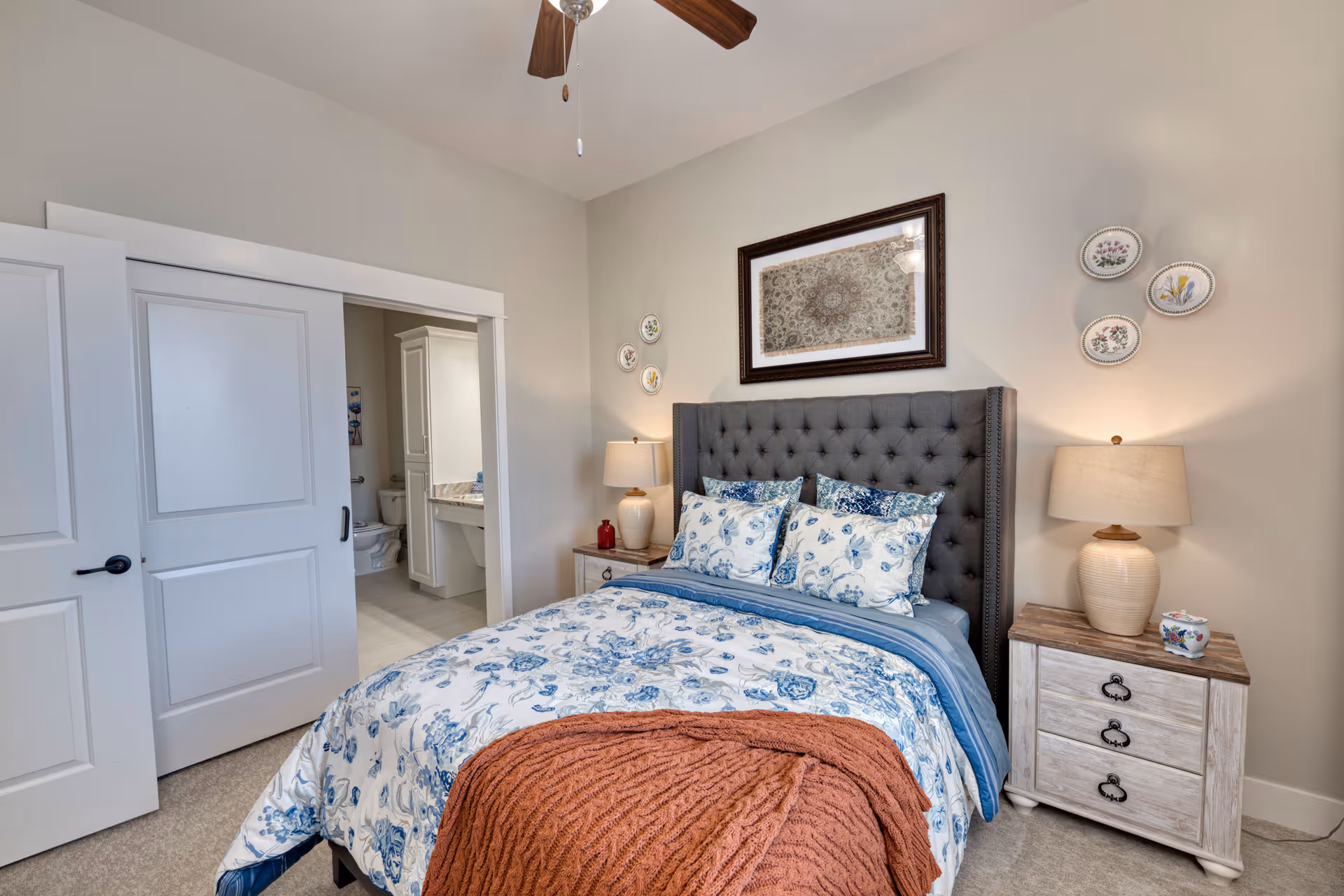 A cozy bedroom with a tufted gray headboard bed dressed in blue and white floral bedding and a rust-colored throw blanket. There are two wooden nightstands on either side of the bed, each with a lamp and decorative items. Above the bed hangs a framed artwork, and three small decorative plates are mounted on the wall to the right. A ceiling fan is visible above, and an open sliding door reveals a bathroom with a toilet and vanity.