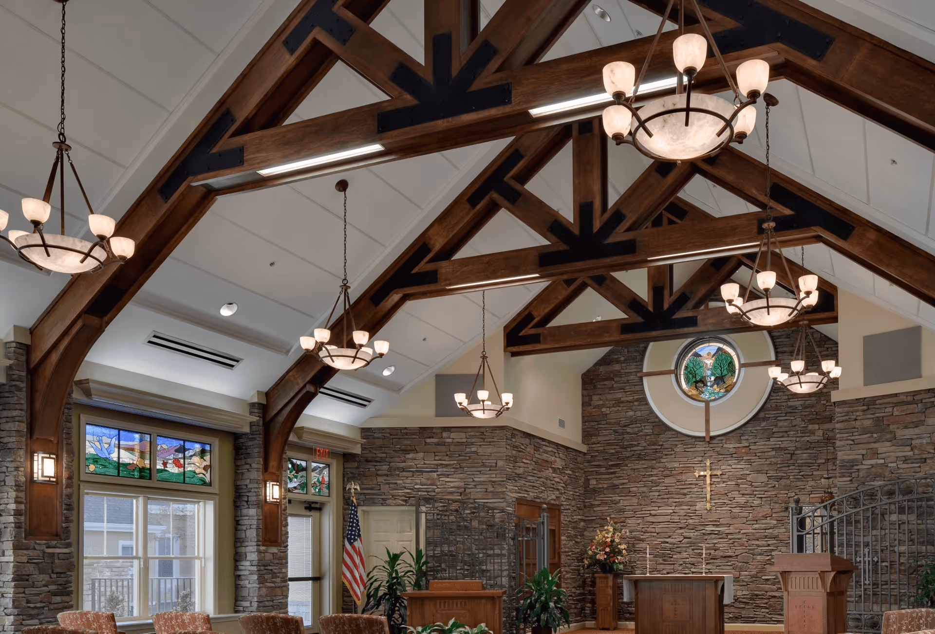 Interior view of a chapel or worship space with high vaulted ceilings supported by dark wooden beams, stone walls, stained glass windows, chandeliers, an altar with a cross on the wall behind it, and an American flag to the side.