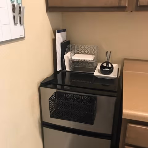 A small stainless steel mini refrigerator with a black decorative basket attached to its front. On top of the refrigerator, there is a clear organizer holding papers and napkins, a small white dish with a black round object and two pens. The refrigerator is placed against a beige wall and next to a brown countertop and wooden cabinets.