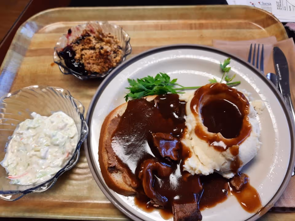 A meal tray with a plate of sliced roast beef covered in brown gravy, mashed potatoes with brown gravy, and a garnish of parsley. On the tray are two small bowls, one with a creamy salad and the other with a fruit crumble dessert.