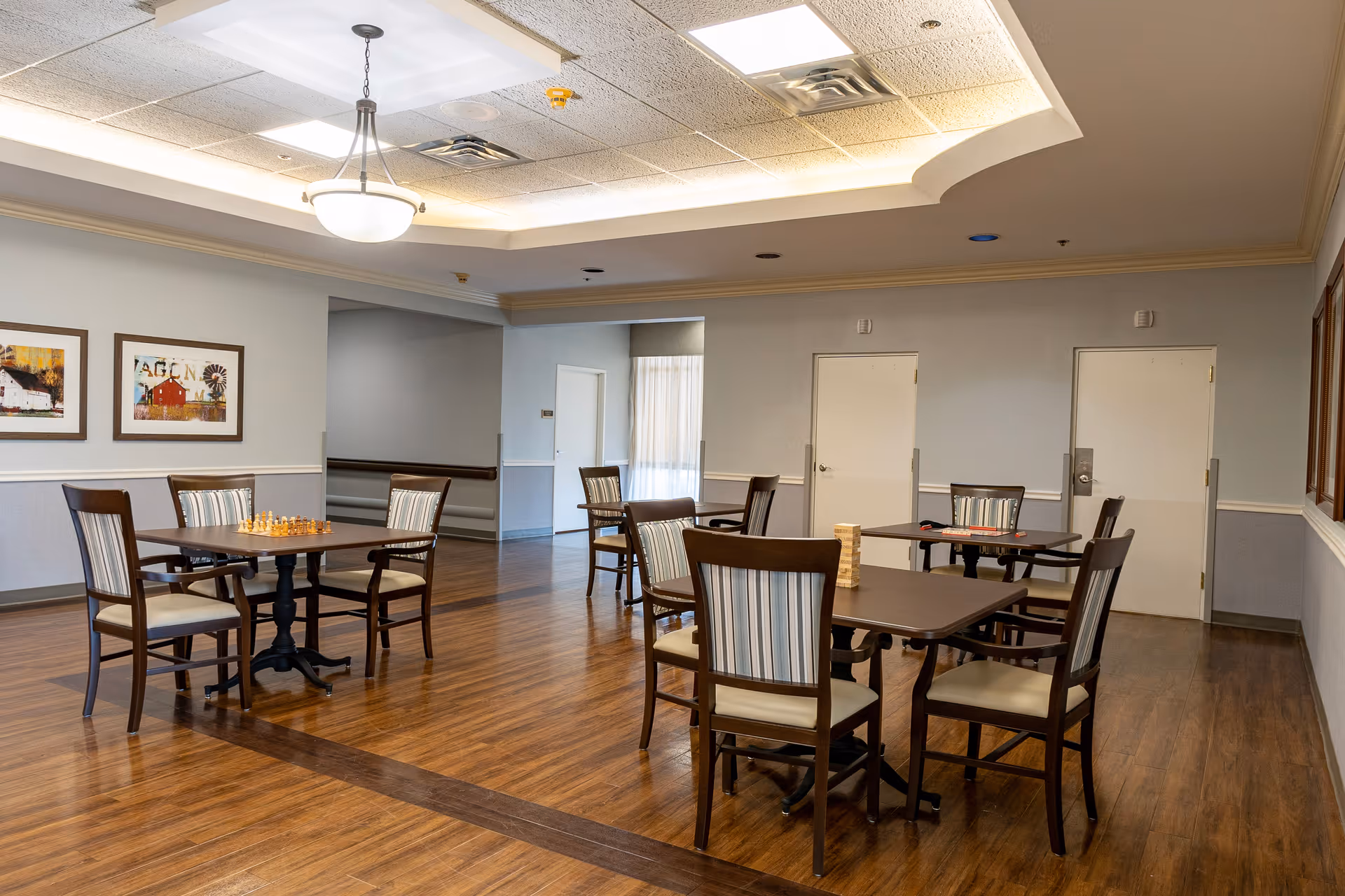 A spacious room with wooden flooring and several tables and chairs arranged for seating. Each table has a board game set up, including chess and Jenga. The walls are light-colored with framed artwork, and the ceiling has recessed lighting and a hanging light fixture.