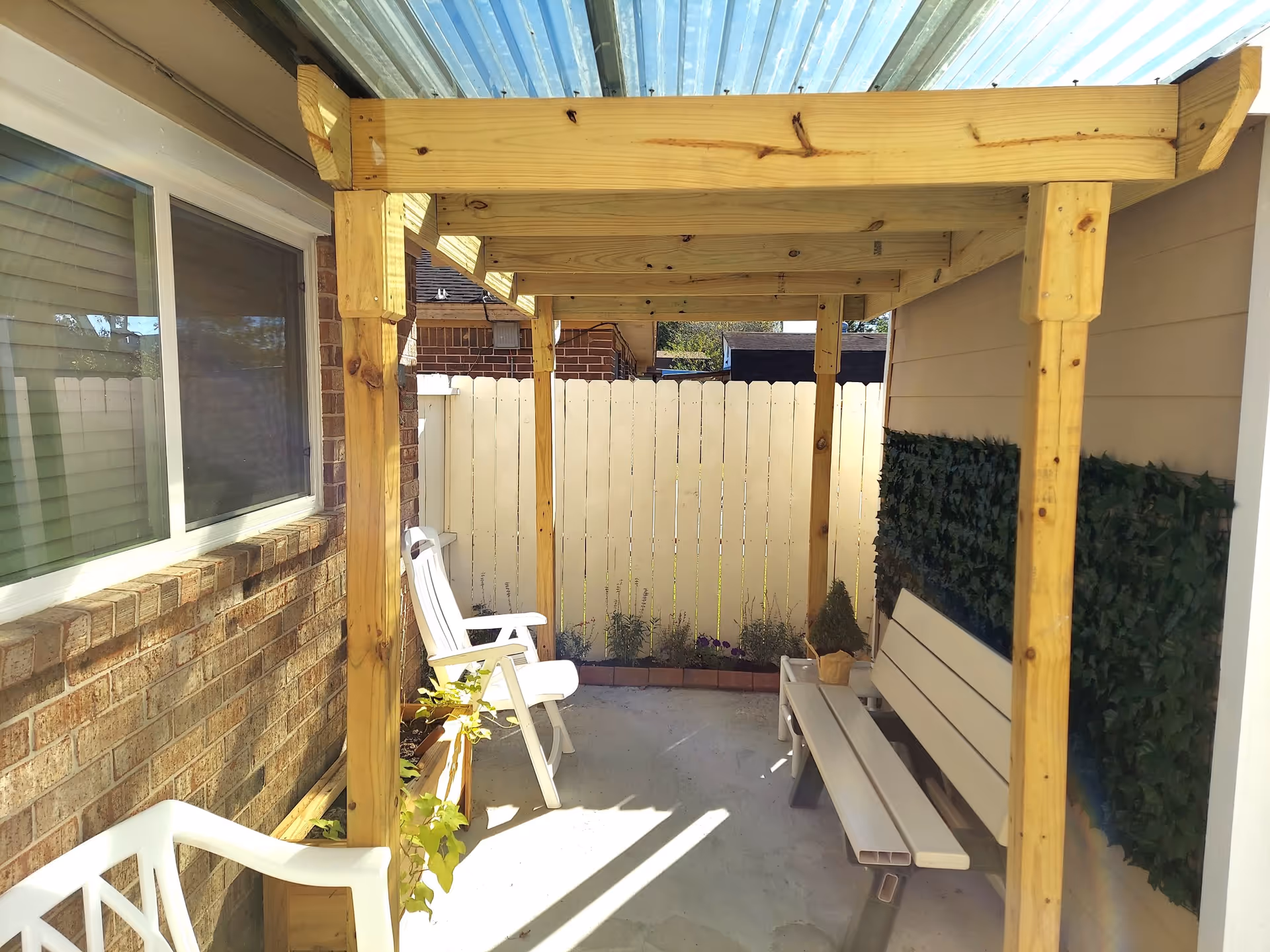 Small covered patio with a wooden pergola, white plastic chairs and a bench beside a brick wall and white fence.