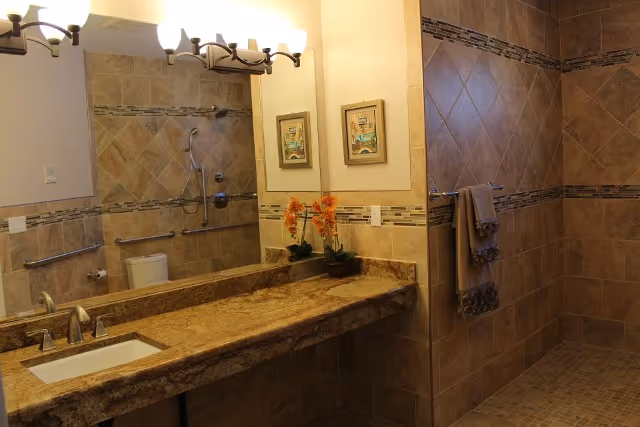 A bathroom with beige and brown tiled walls and floor. It features a granite countertop with a built-in sink, a large mirror above the sink, two framed pictures on the wall, and a light fixture with multiple bulbs. There is a walk-in shower with a towel hanging on a bar and a grab bar installed on the wall.