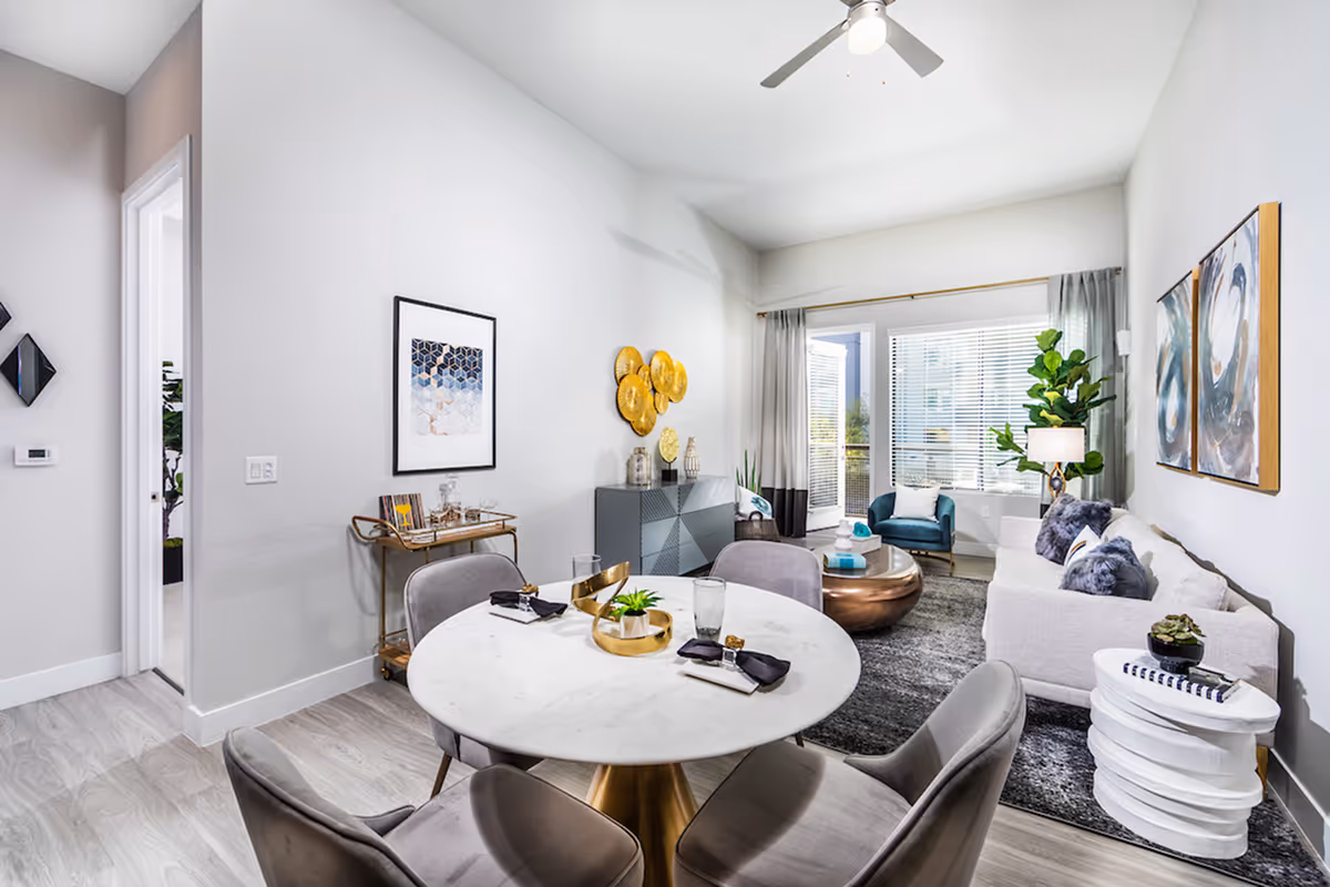 A modern living and dining area with a round white marble dining table surrounded by four gray chairs. The living room features a white sofa with gray pillows, a round copper coffee table, a blue armchair, a large window with blinds, and a tall green plant. The walls are decorated with abstract art and gold circular wall decor. Light wood flooring and a ceiling fan complete the space.