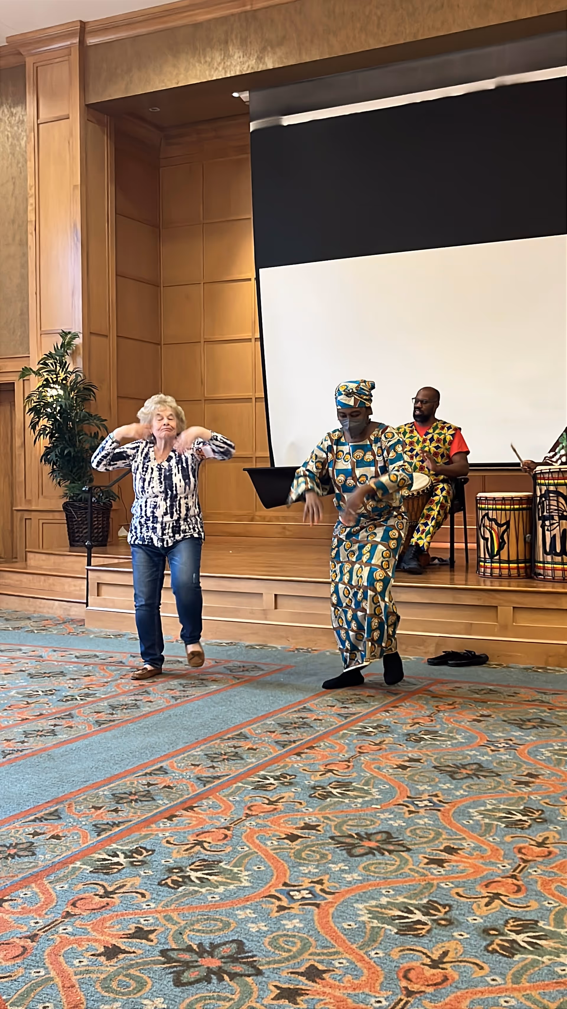 An elderly woman and a person dressed in colorful traditional African attire are dancing in a room with a patterned carpet and wooden panel walls. Behind them, a man in matching colorful attire is seated on a stage playing a drum. There is a large white screen on the stage and a potted plant to the left.