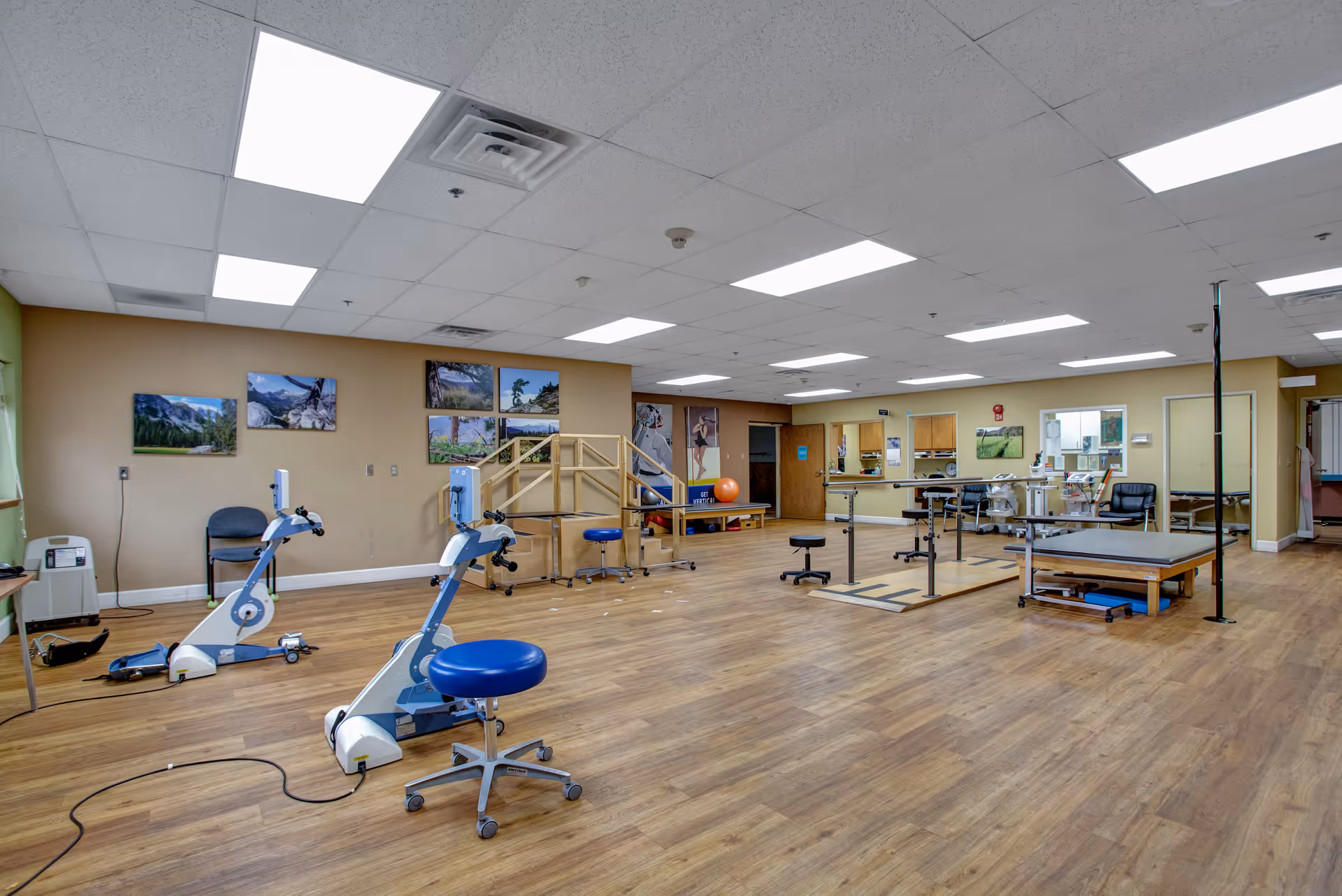 A spacious physical therapy or rehabilitation room with wooden flooring, two stationary exercise bikes, parallel bars for walking exercises, a padded therapy table, stools, and various exercise equipment. The walls are decorated with landscape photos, and the ceiling has multiple fluorescent lights.