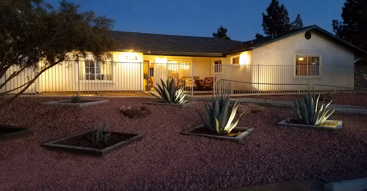 Exterior view of a single-story building at dusk with warm lights illuminating the porch area. The building has a ramp with railings leading to the entrance. The foreground features a landscaped area with gravel and several agave plants in square concrete planters.