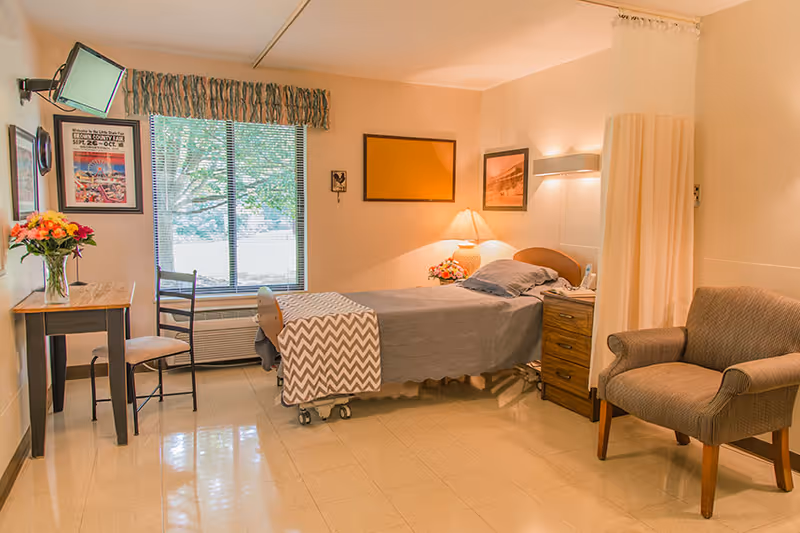 A well-lit rehabilitation room with a single hospital bed covered with a gray blanket and a chevron-patterned throw. There is a wooden nightstand with a lamp and flowers next to the bed, a comfortable armchair, a small table with a chair and a vase of flowers, a wall-mounted TV, and a window with blinds and a valance. The room has light-colored walls and a privacy curtain partially drawn.