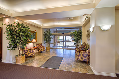 A spacious and well-lit lobby area with tiled flooring and a carpeted section. There are four patterned armchairs arranged in two groups, two on each side, with side tables and lamps. Two large potted plants flank the entrance doors, which are glass and lead outside. The walls are painted light beige, and there are modern wall sconces providing additional lighting.