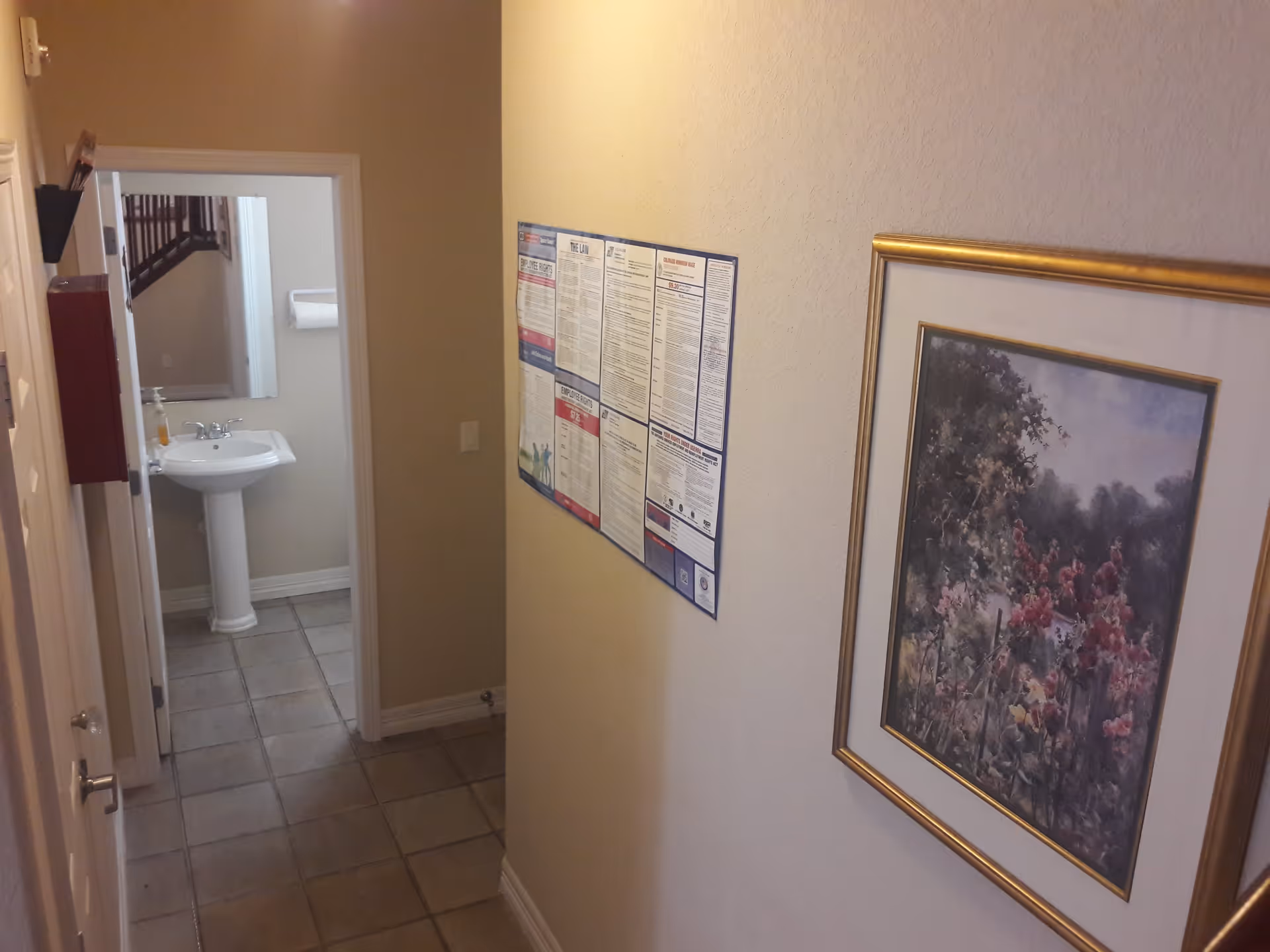 A hallway with beige walls and tiled floor leading to a bathroom. The bathroom has a white pedestal sink, a mirror, and a paper towel dispenser. On the hallway wall, there is a framed floral painting and a bulletin board with various notices.