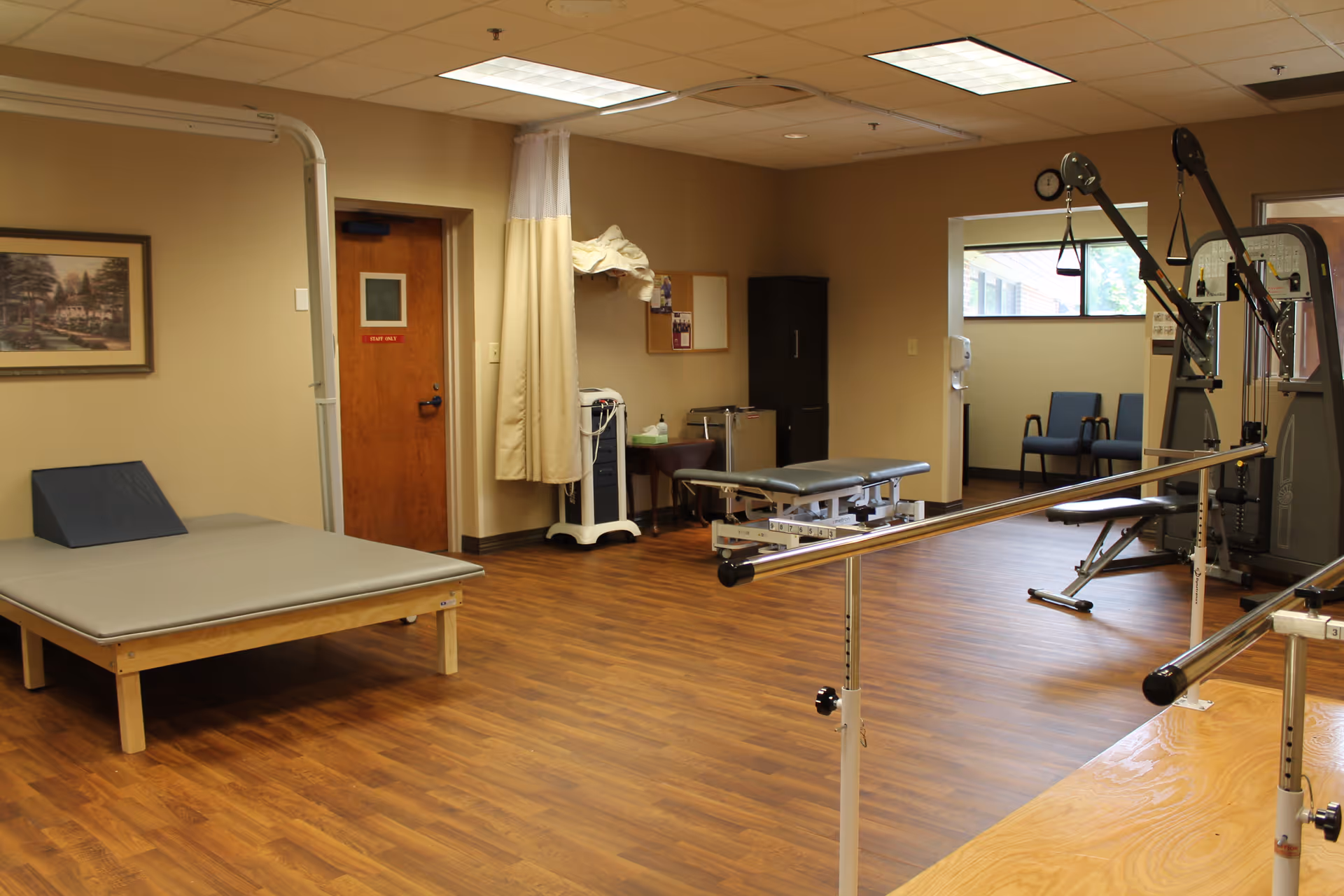 A rehabilitation room with wooden flooring featuring parallel bars for walking exercises, two padded treatment tables, exercise equipment, a wooden door, a framed picture on the wall, and a small seating area with two chairs near a window.
