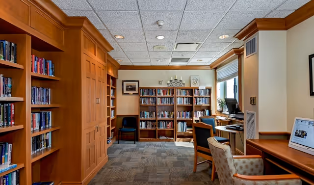 A cozy library or reading room with wooden bookshelves filled with books, a few chairs, a computer workstation by the window, and soft overhead lighting. The room has a carpeted floor and a ceiling with recessed lights.