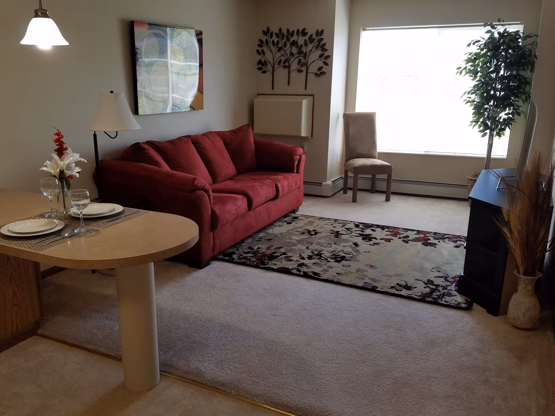 Cozy living room with a red sofa, patterned rug, dining counter with place settings, and a large window with a chair.